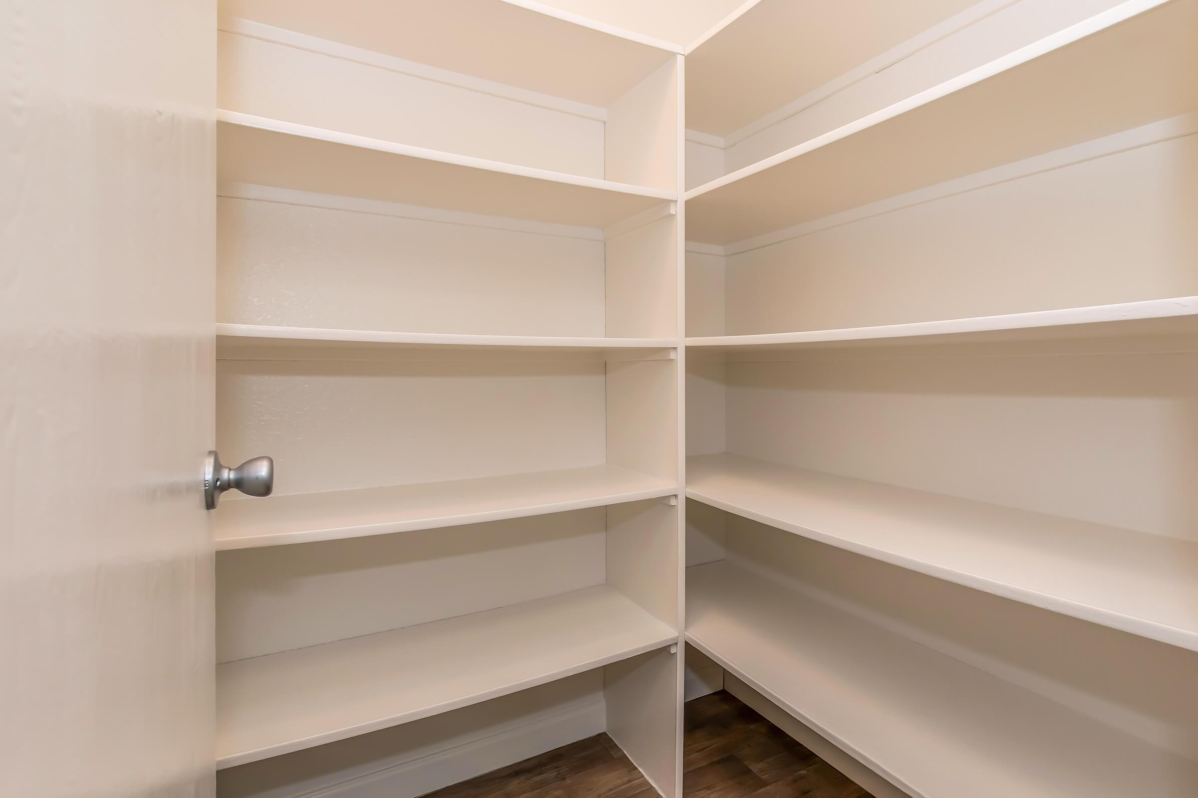 A clean and empty pantry with several white shelves. The walls are painted a light color, and the floor features a wood-like finish. The room is well-lit and has a door that is partially open.