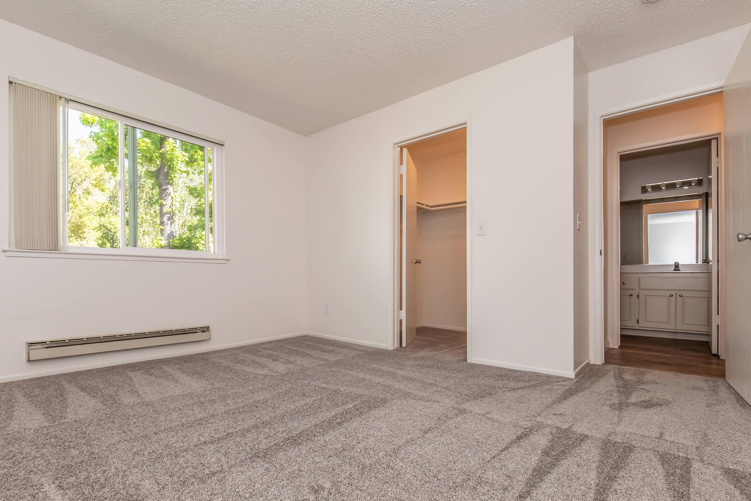 A spacious, empty room with light-colored walls and grey carpet. A window on the left lets in natural light, while a doorway on the right leads to a bathroom. There is a closet visible in the adjacent room. The overall atmosphere is bright and clean, suited for a cozy living space.
