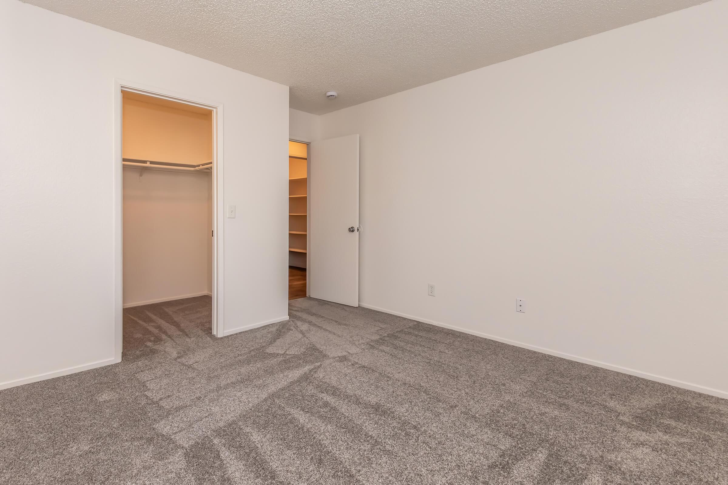 A vacant room with light-colored walls and carpet. The room has an open doorway leading to a closet on one side and another closed door on the opposite side. The lighting is soft, and the space is tidy and uncluttered, suggesting a fresh or recently renovated area.