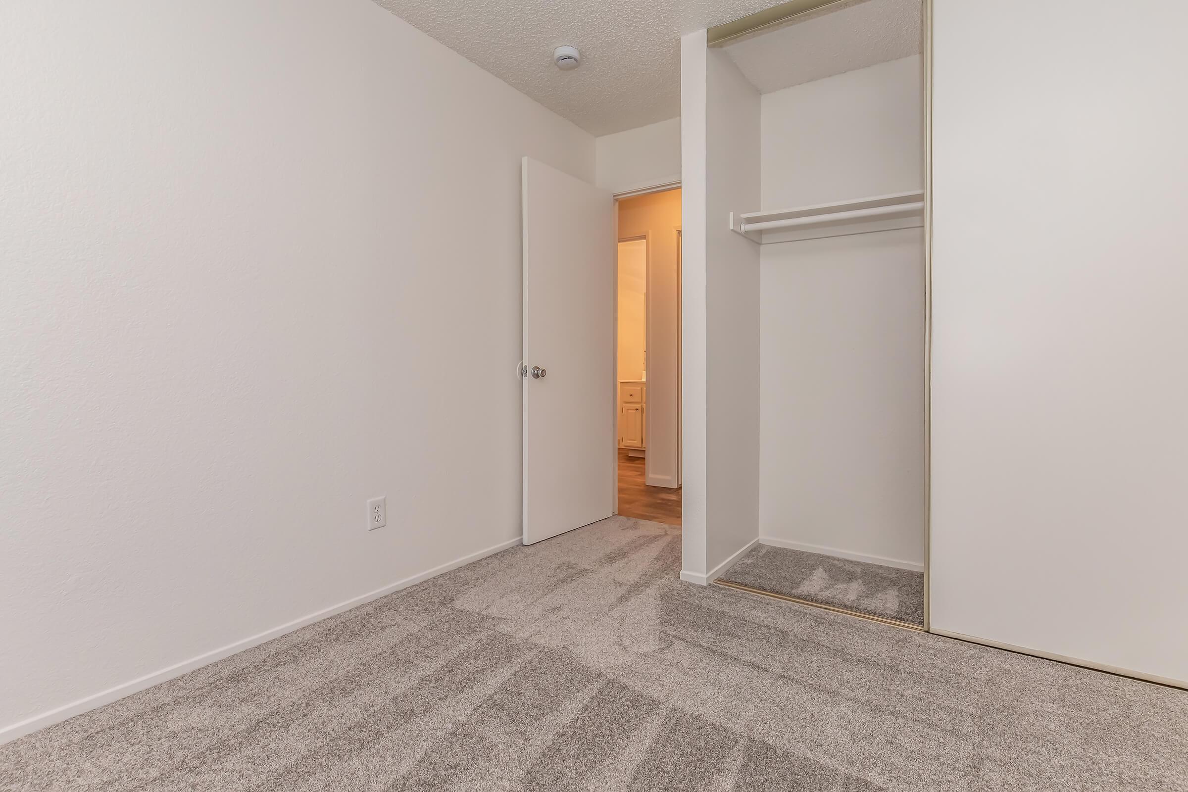 A small, empty room with light-colored walls and carpet. It features an open closet with a hanging rod and a door leading to a bathroom area visible in the background. The overall ambiance is clean and minimalistic, with soft lighting illuminating the space.