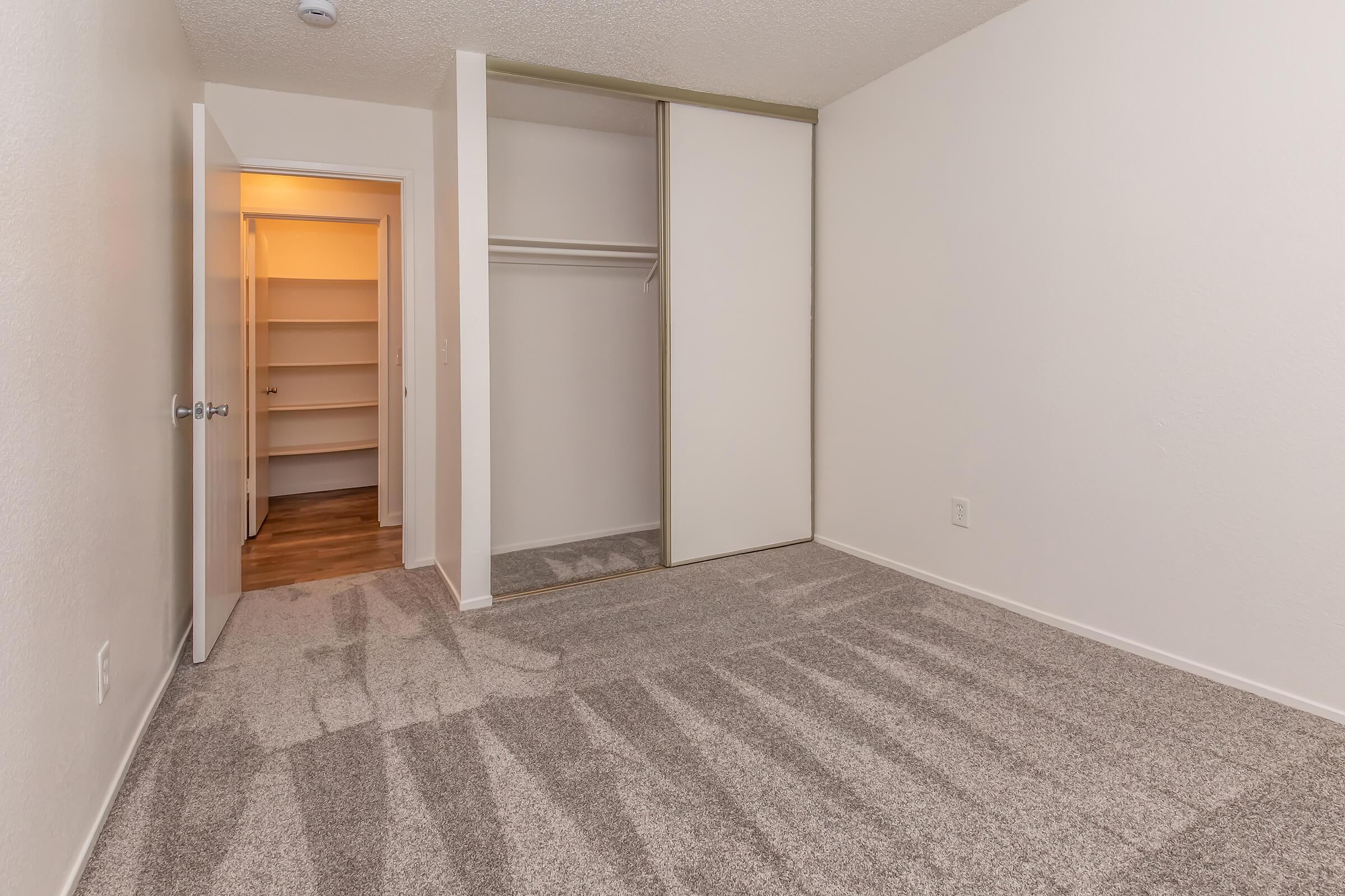 Empty room with light-colored walls and carpeted flooring. Contains a closet with sliding doors on one side and a doorway leading to a storage area on the left. Natural light enters from the open door, creating a bright, clean appearance. The space is unfurnished and ready for personal decoration.