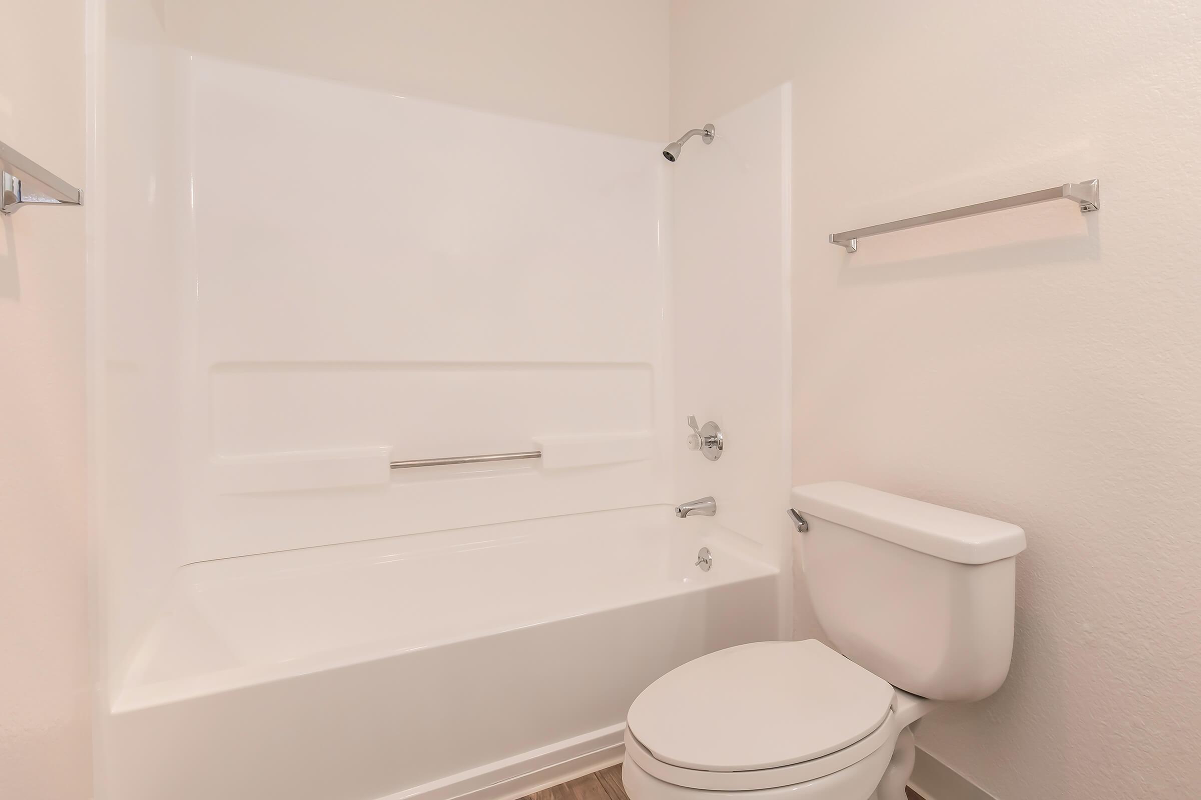 A clean, modern bathroom featuring a white shower/tub combination with a grab bar, a sleek toilet, and light-colored walls. The space has an overall minimalist design with no decorative elements.