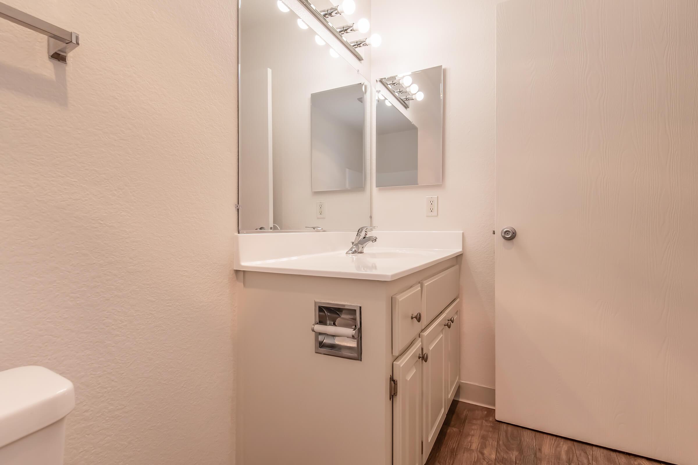 A modern bathroom featuring a white vanity with a sink and mirror, illuminated by bright light fixtures. The walls are painted a light color, and a door partially closed is visible on the right. A portion of a toilet is seen in the lower left corner. The flooring is a light wood.