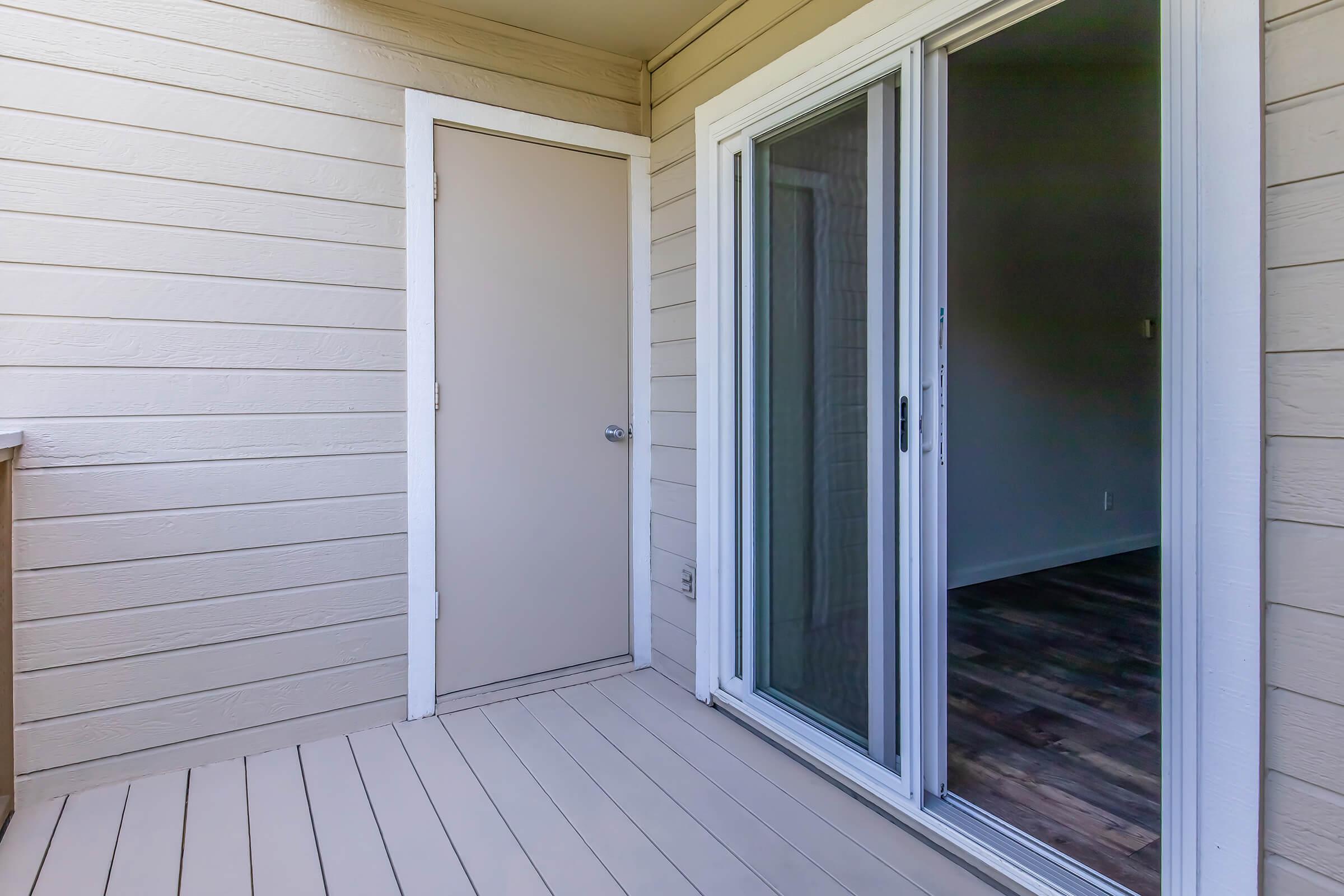 A view of a small balcony or patio featuring a sliding glass door that leads into an indoor space. The walls are made of light-colored wood siding, and there is a plain door on the left side. The floor is a light-colored composite material, adding a modern touch to the area.