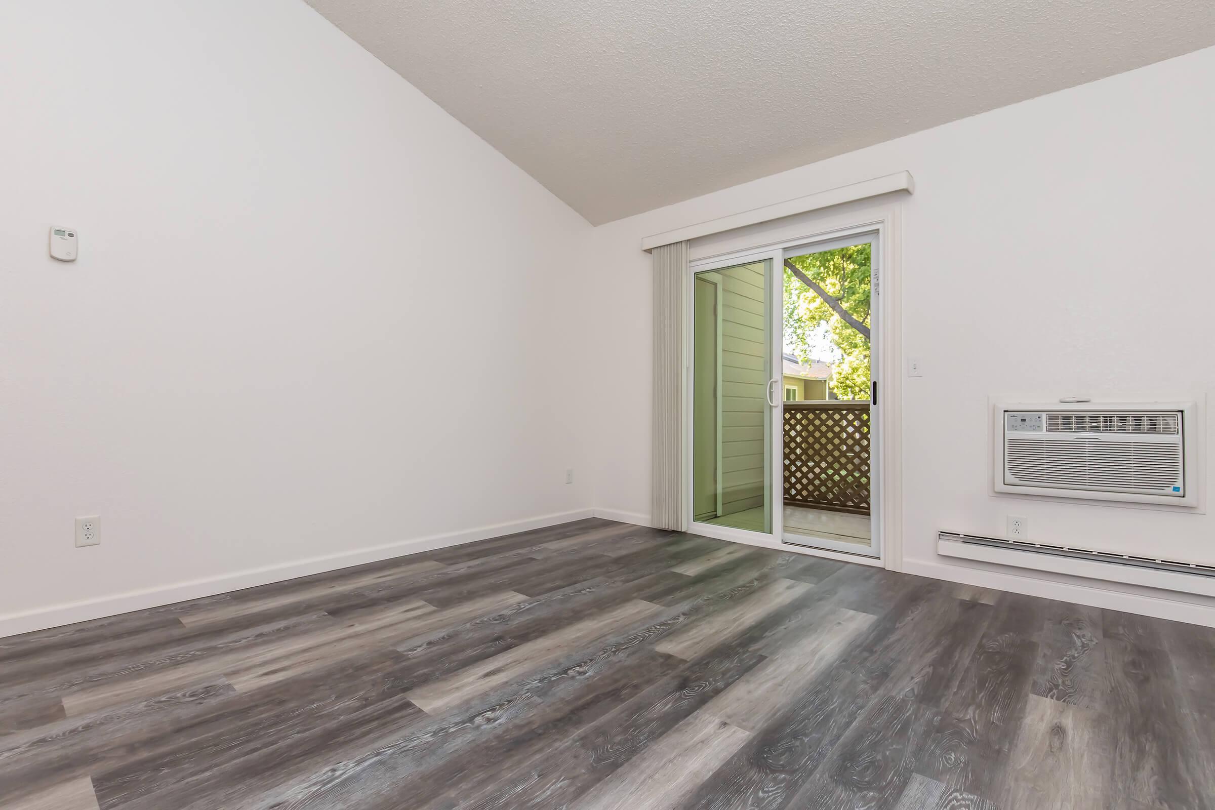 A spacious, unfurnished room with a high ceiling and large windows. The floor features modern gray laminate, and there is a sliding glass door leading to a small balcony. An air conditioning unit is mounted on the wall, and the walls are painted white, creating a bright and airy atmosphere.
