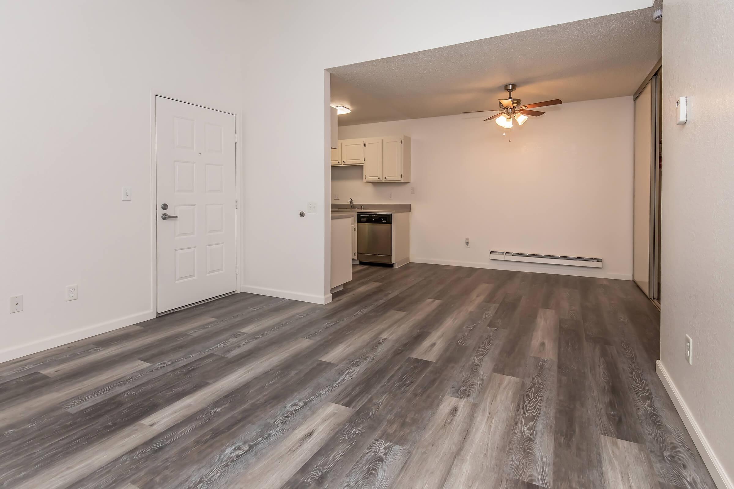 An empty room featuring light-colored walls and grey laminate flooring. A ceiling fan hangs in the corner, and a doorway leads to a kitchen area with white cabinetry and a countertop. Natural light brightens the space, emphasizing its spaciousness and simplicity.