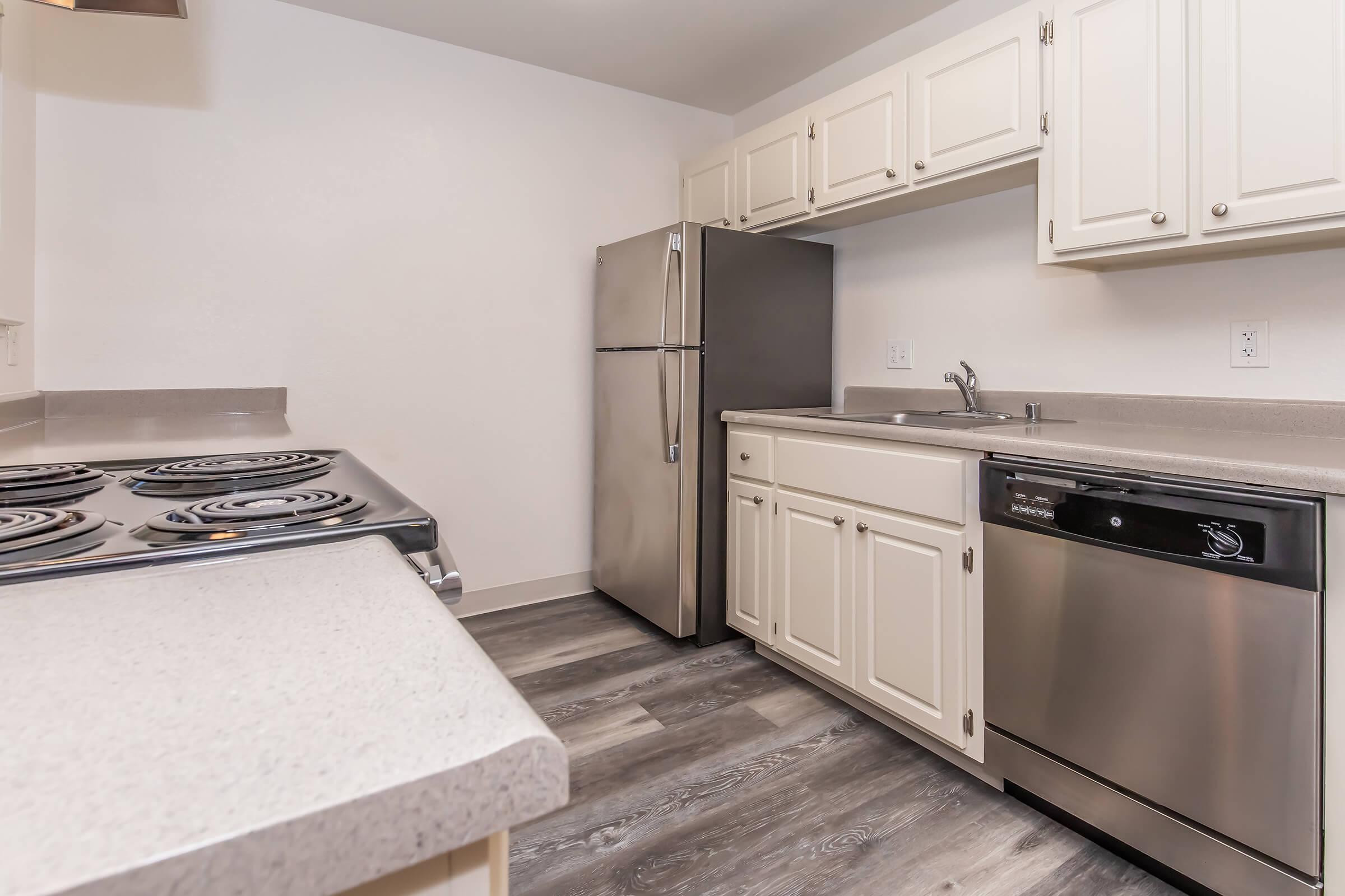 A modern kitchen featuring beige cabinetry, stainless steel appliances including a refrigerator and dishwasher, a ceramic cooktop, and gray countertop surfaces. The flooring is a light-toned laminate, creating a clean and contemporary look. Natural light fills the space.