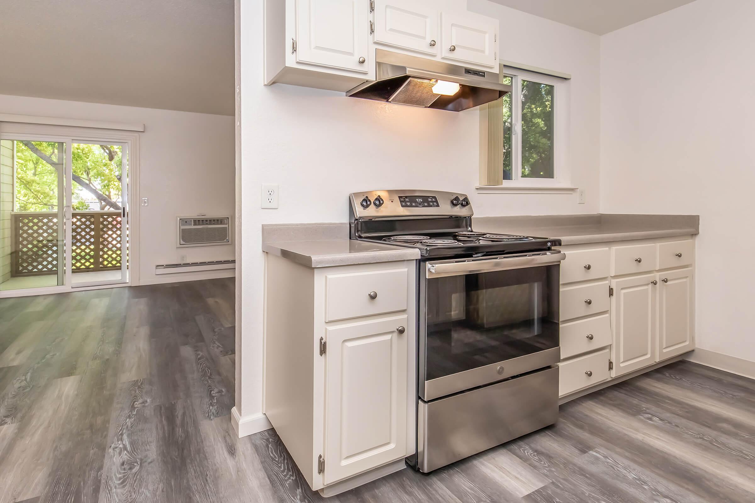 Kitchen interior featuring a stainless steel gas stove with an overhead exhaust fan, white cabinetry, and a countertop. The space has a modern design with a window providing natural light, and adjacent to the kitchen, a sliding door leads to an outdoor balcony area. The flooring is a light-colored laminate.
