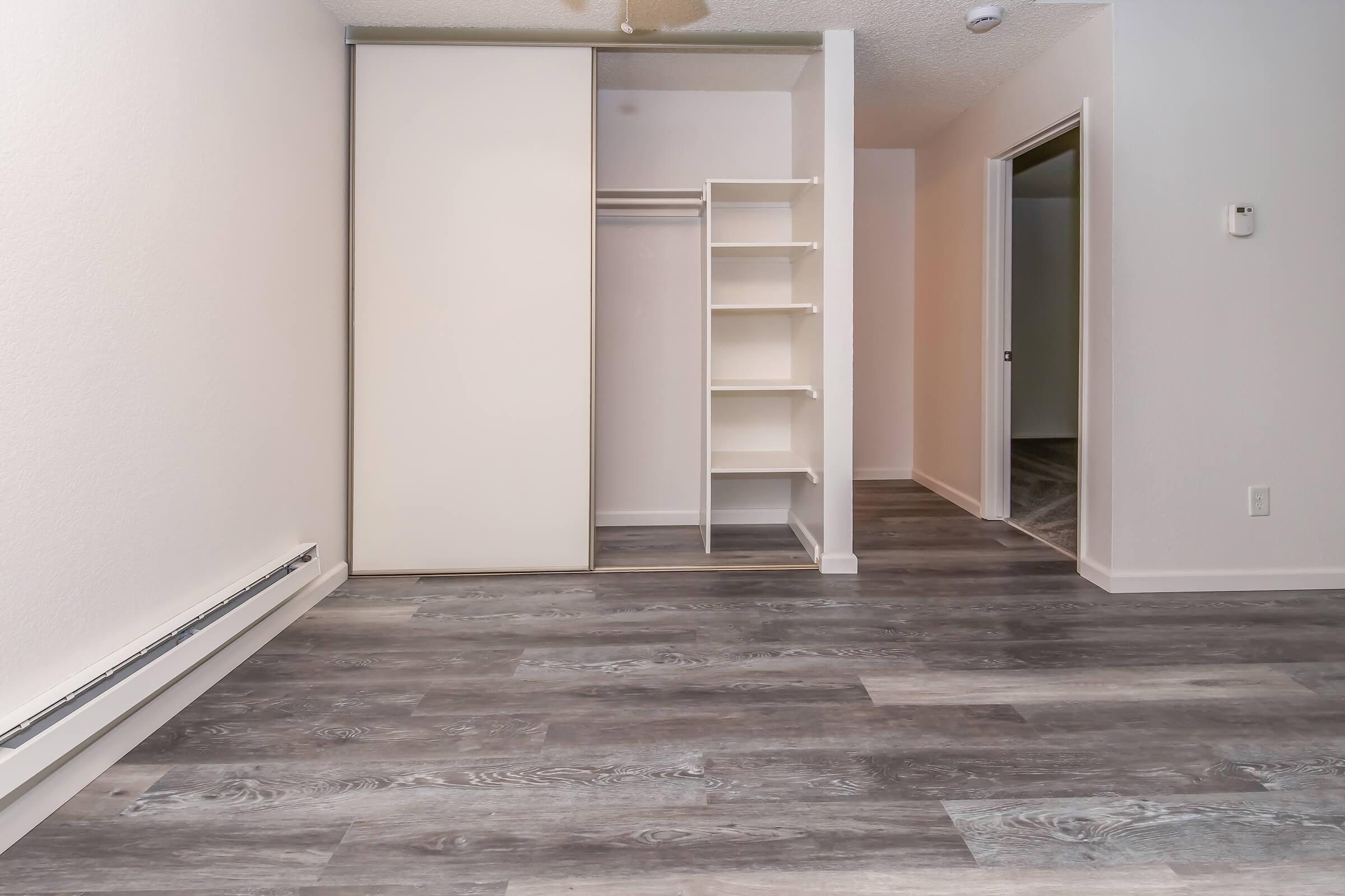 Empty interior of a room featuring light-colored walls and a gray laminate floor. There is a sliding closet door on the left, with a shelf unit beside it. A door leads to another room in the background, and a ceiling fan is visible above. The space appears bright and clean, suitable for various furnishings.