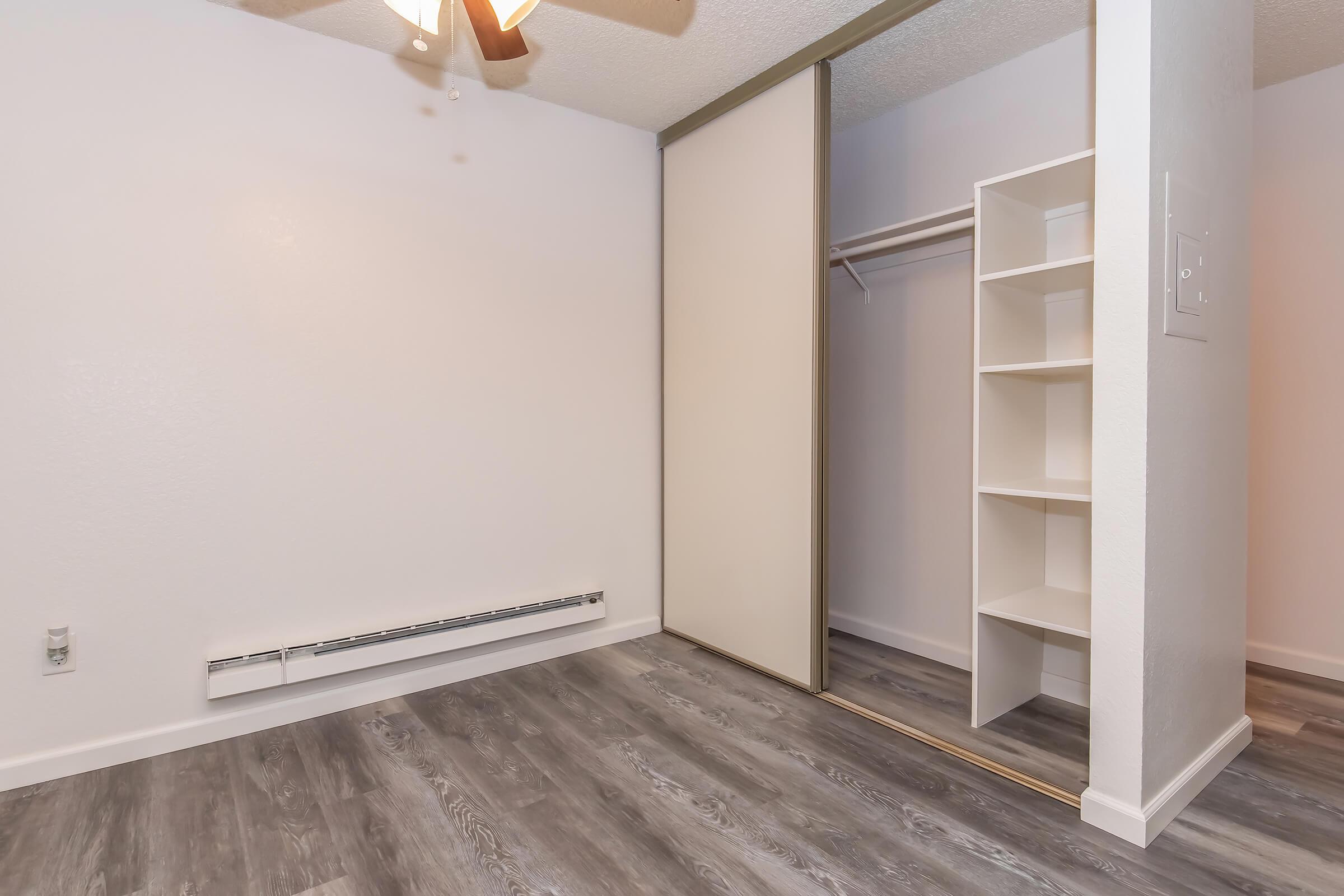 Empty room featuring light-colored walls, a ceiling fan, and a closet with sliding doors. The floor has a tan-colored laminate, and there is a heating unit along the wall. The closet has built-in shelving and a hanging rod. The overall atmosphere is bright and minimalist.