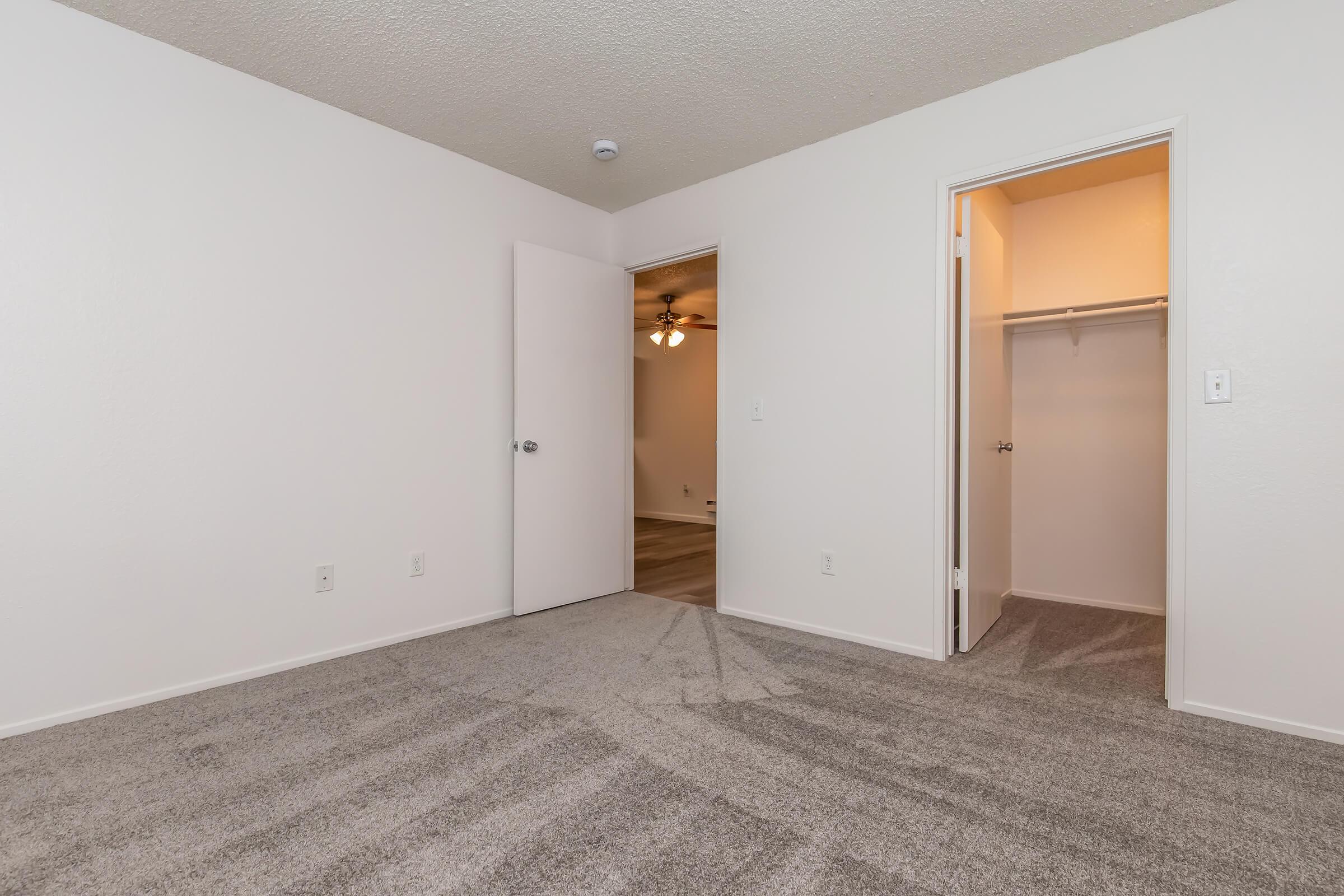 A spacious interior of an empty room featuring light gray carpet, white walls, and a closed door leading to a closet. An adjacent door opens to another room with a ceiling fan visible. The overall ambiance is bright and inviting, suitable for personalization.