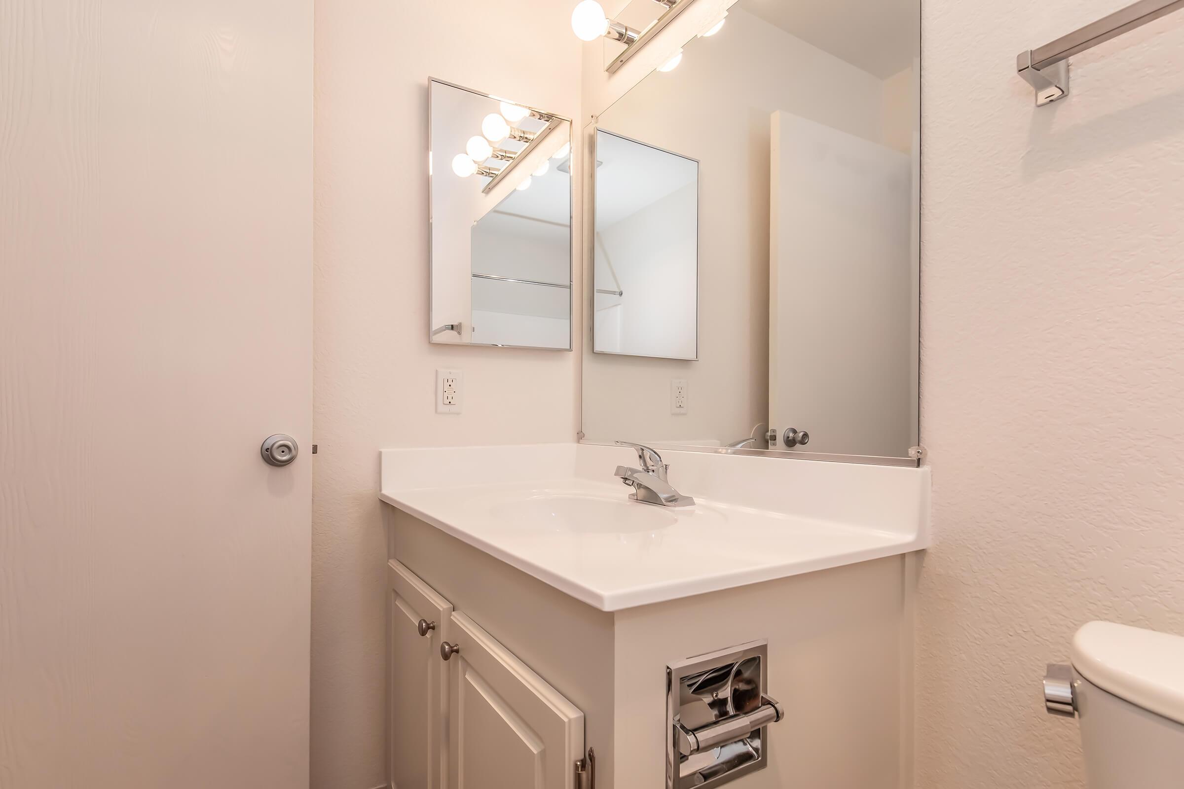 A clean and modern bathroom featuring a white vanity with a sink, a large mirror above, and bright light fixtures. The walls are a light color, and there is a door partially visible on the left side. A towel holder is mounted on the wall beside the toilet, contributing to the tidy and minimalist aesthetic.