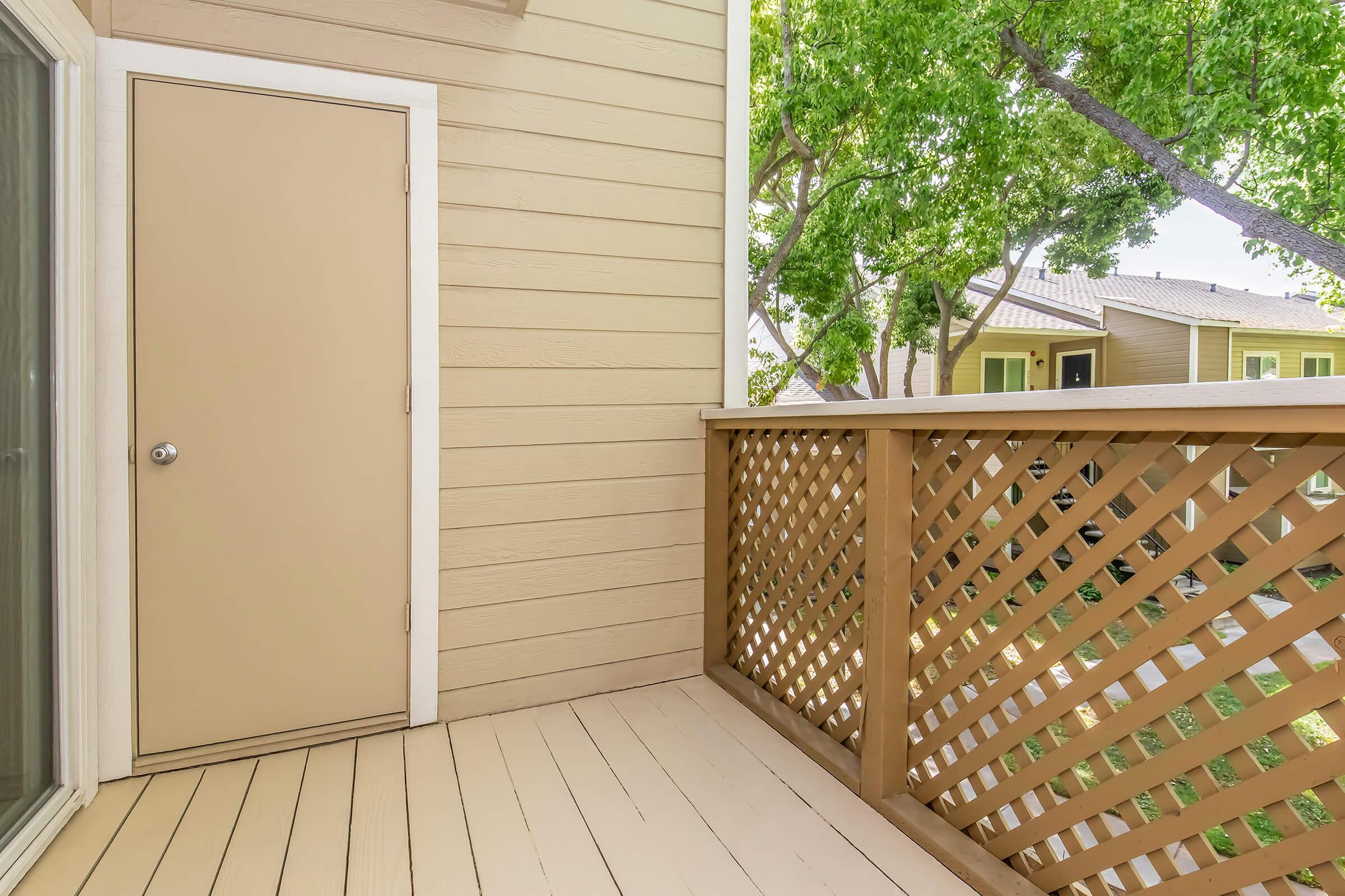 A small balcony with wood lattice railing, featuring a closed door on one side. The flooring is light-colored and there are trees visible nearby, providing a green backdrop to the outdoor space.