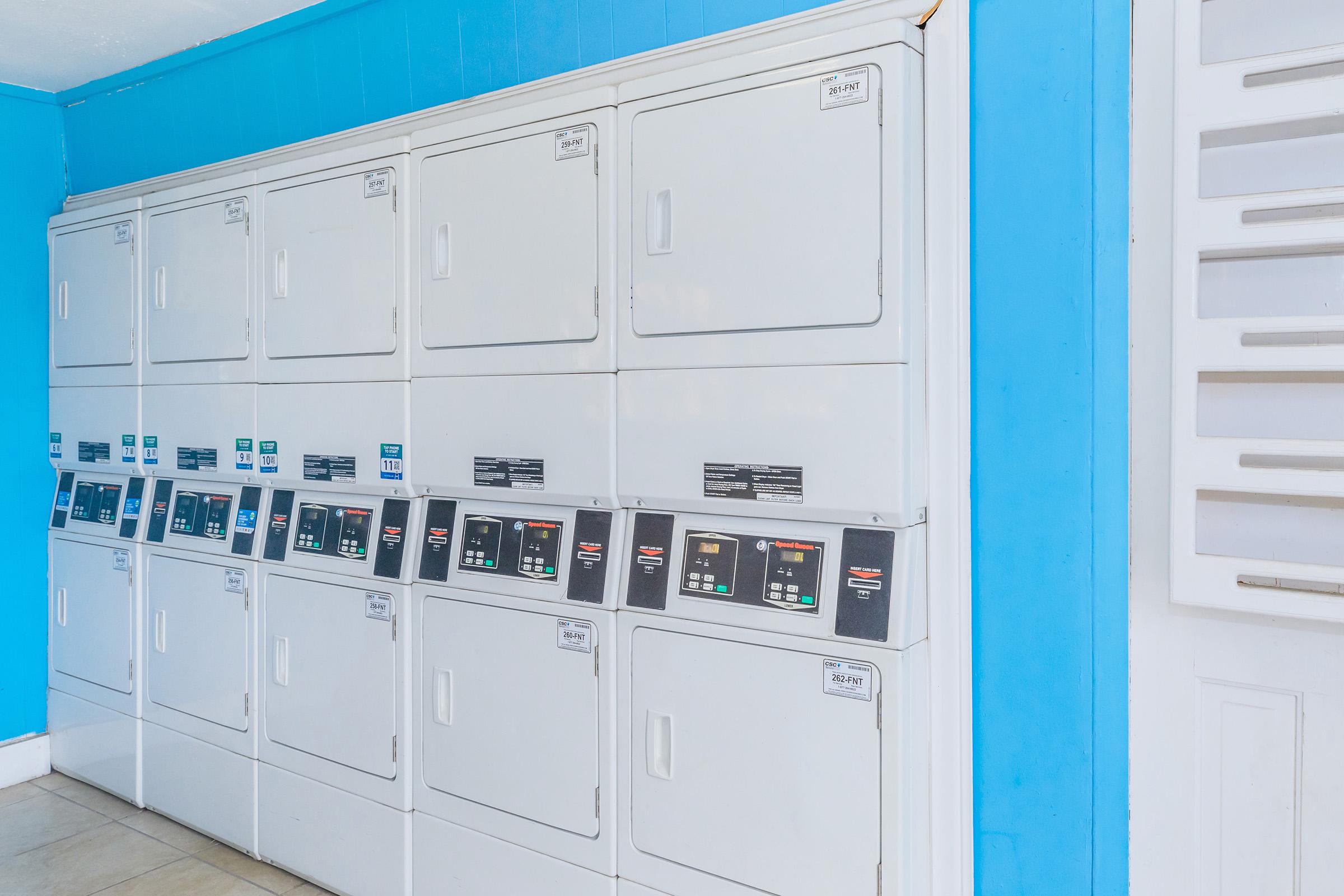 A row of white stacked laundry machines against a light blue wall. The machines feature control panels with buttons and digital displays. The floor is tiled, and there’s a white door on the right side of the image.