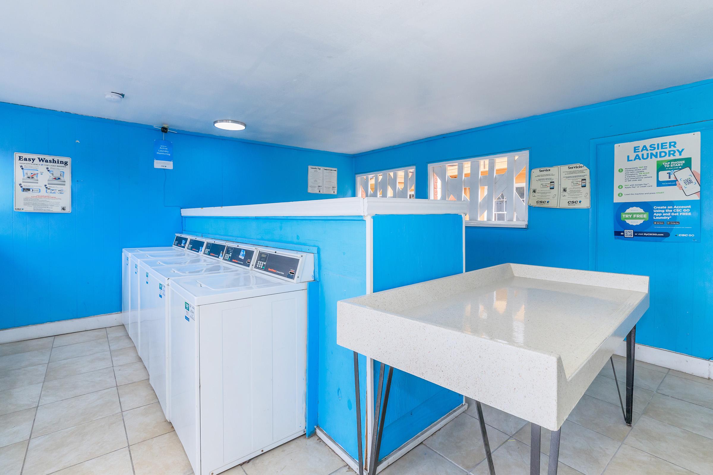 Brightly colored laundry room featuring several white washing machines, a large white folding table, and blue walls. There are informational posters on the walls and windows allowing natural light to enter, creating a clean and inviting atmosphere for laundry activities.