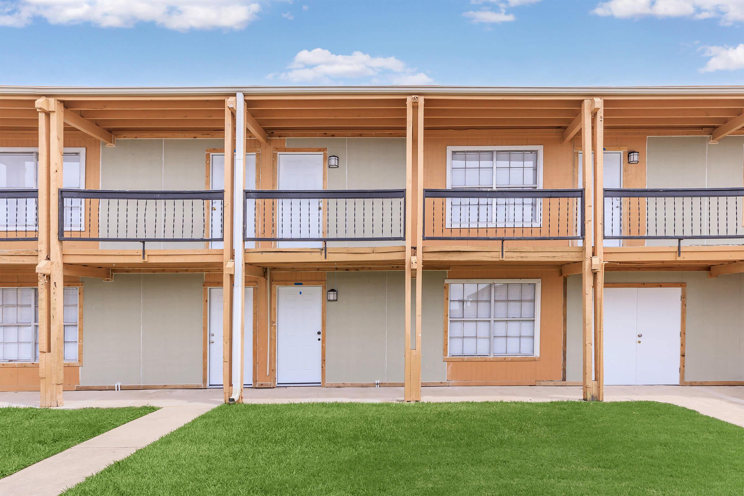 Two-story building with a row of balcony-access apartments. Each unit has a white door and large windows with white frames. The structure features wooden supports and a light brown color scheme. A well-manicured green lawn is in front, with a concrete walkway leading to the doors. The sky is bright blue with a few clouds.