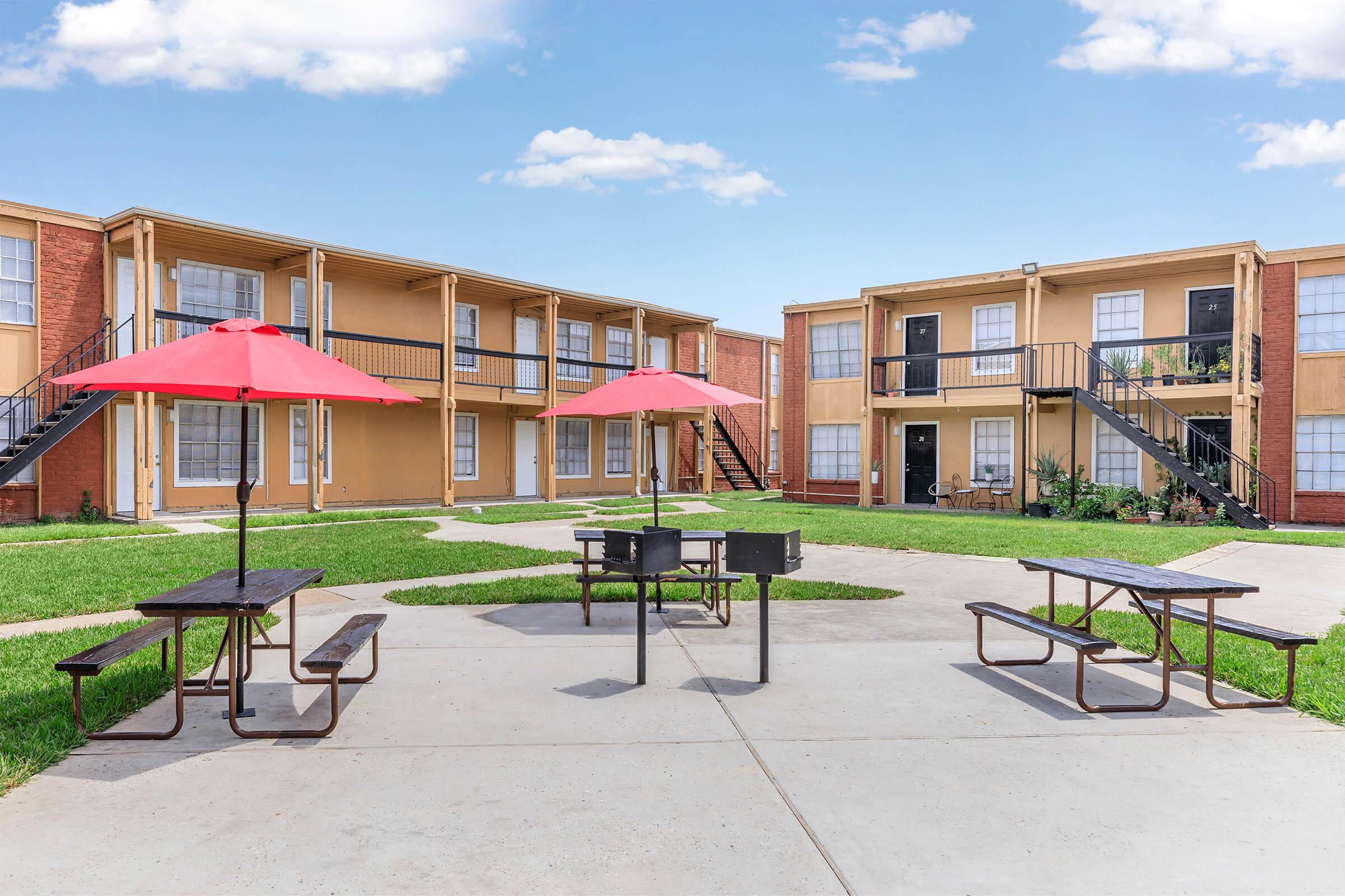 A courtyard area featuring two buildings with balconies, surrounded by green grass. In the center, there are two picnic tables with red umbrellas and a grill, providing a communal outdoor space for residents. The sky is clear with a few clouds, creating a pleasant atmosphere.