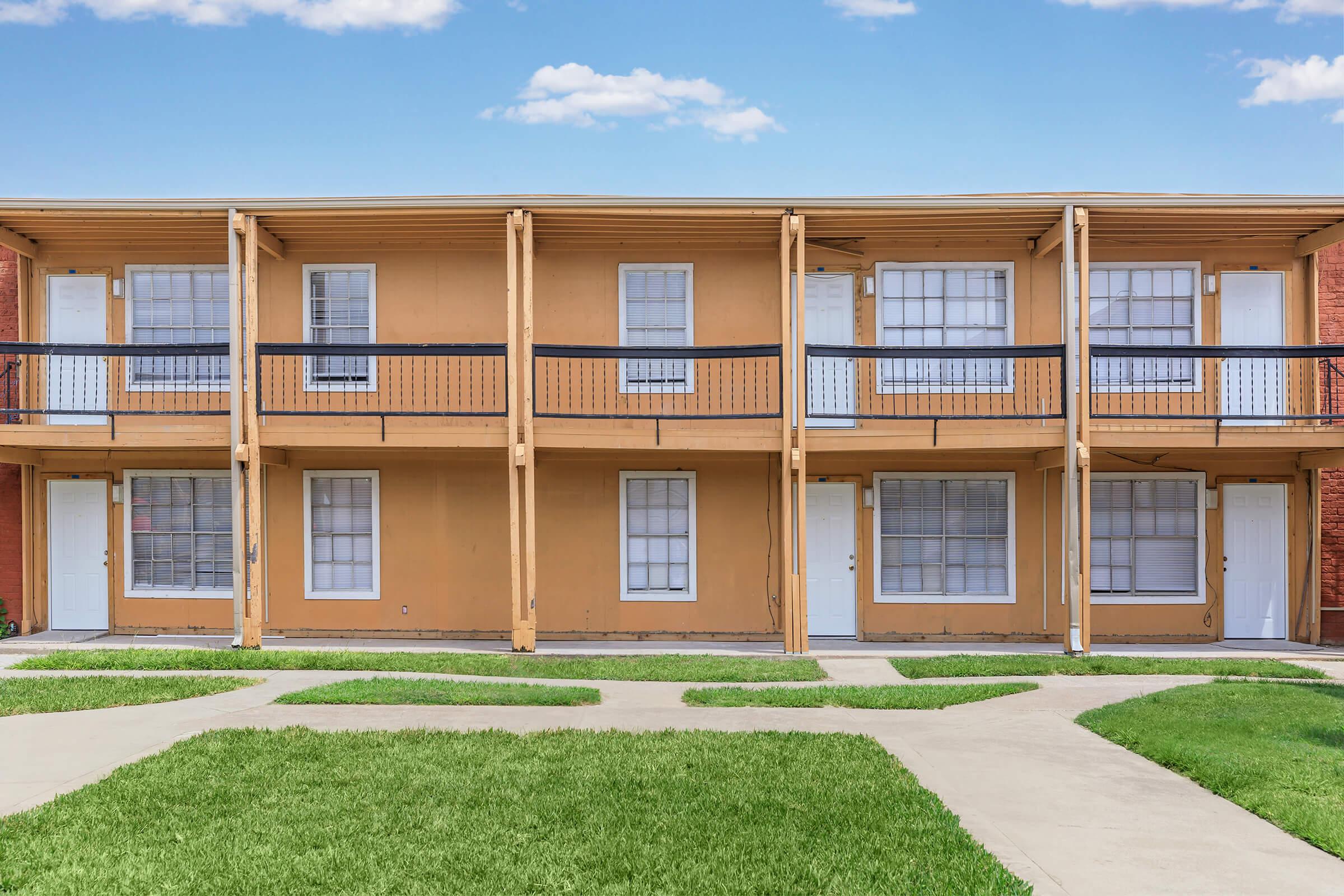 A two-story apartment building with a warm orange exterior. Each unit has a balcony with black railings and white doors. The building is surrounded by a landscaped lawn and a concrete pathway running through the grassy area. The sky above is partly cloudy, creating a bright atmosphere.