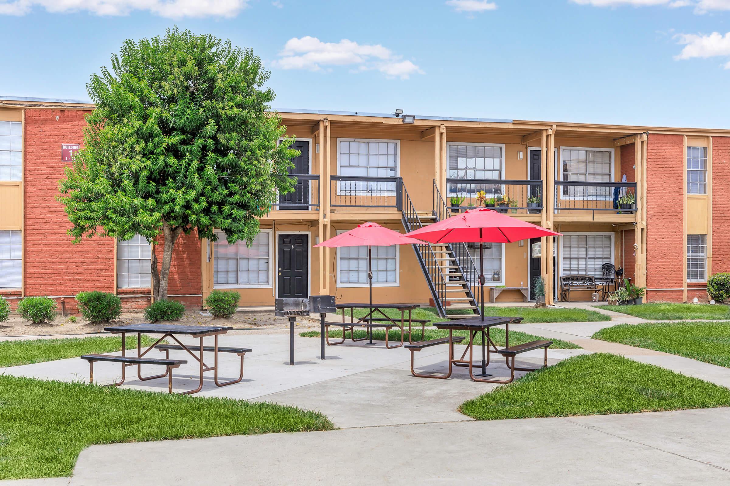 A landscaped courtyard featuring several picnic tables with red umbrellas, surrounded by green grass. In the background, a two-story apartment building with a mix of red and beige siding, large windows, and a staircase leading to the upper units.