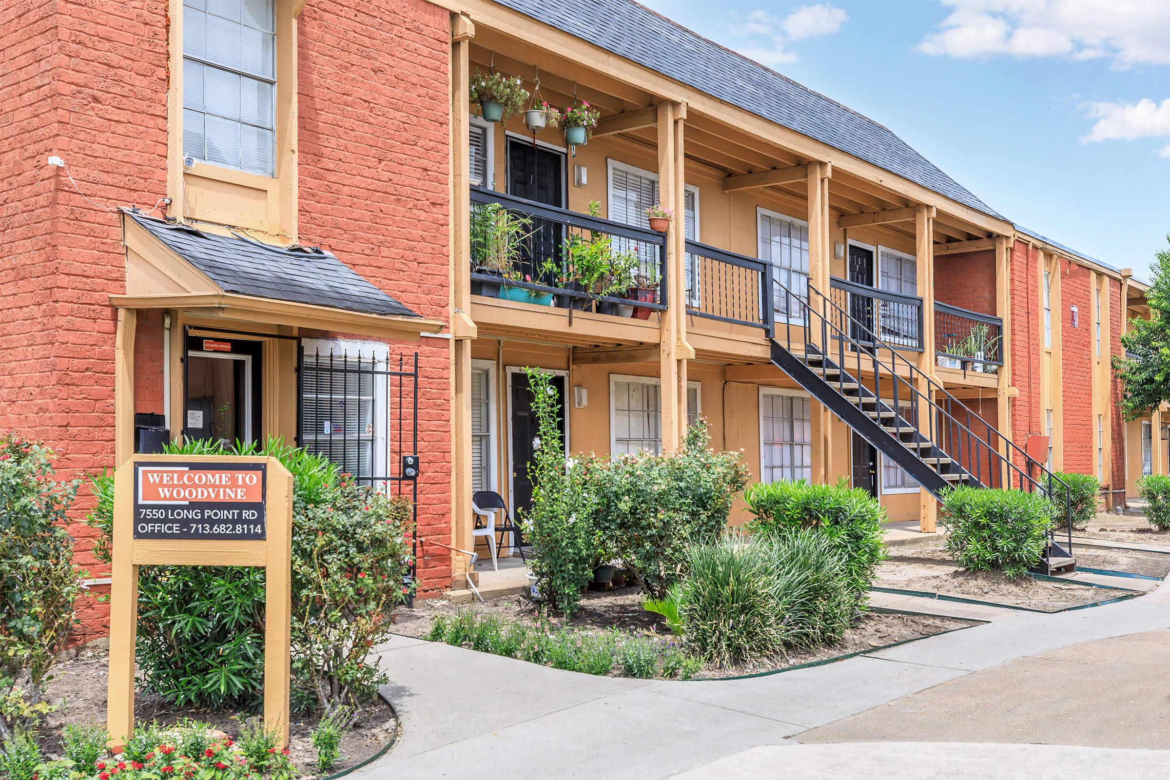 Exterior view of an apartment complex featuring two-story buildings with reddish brick and wooden balconies. Lush green landscaping surrounds the entrance, which has a sign saying "Welcome to Woode." A staircase leads to the upper level, and a pathway offers access to the units.
