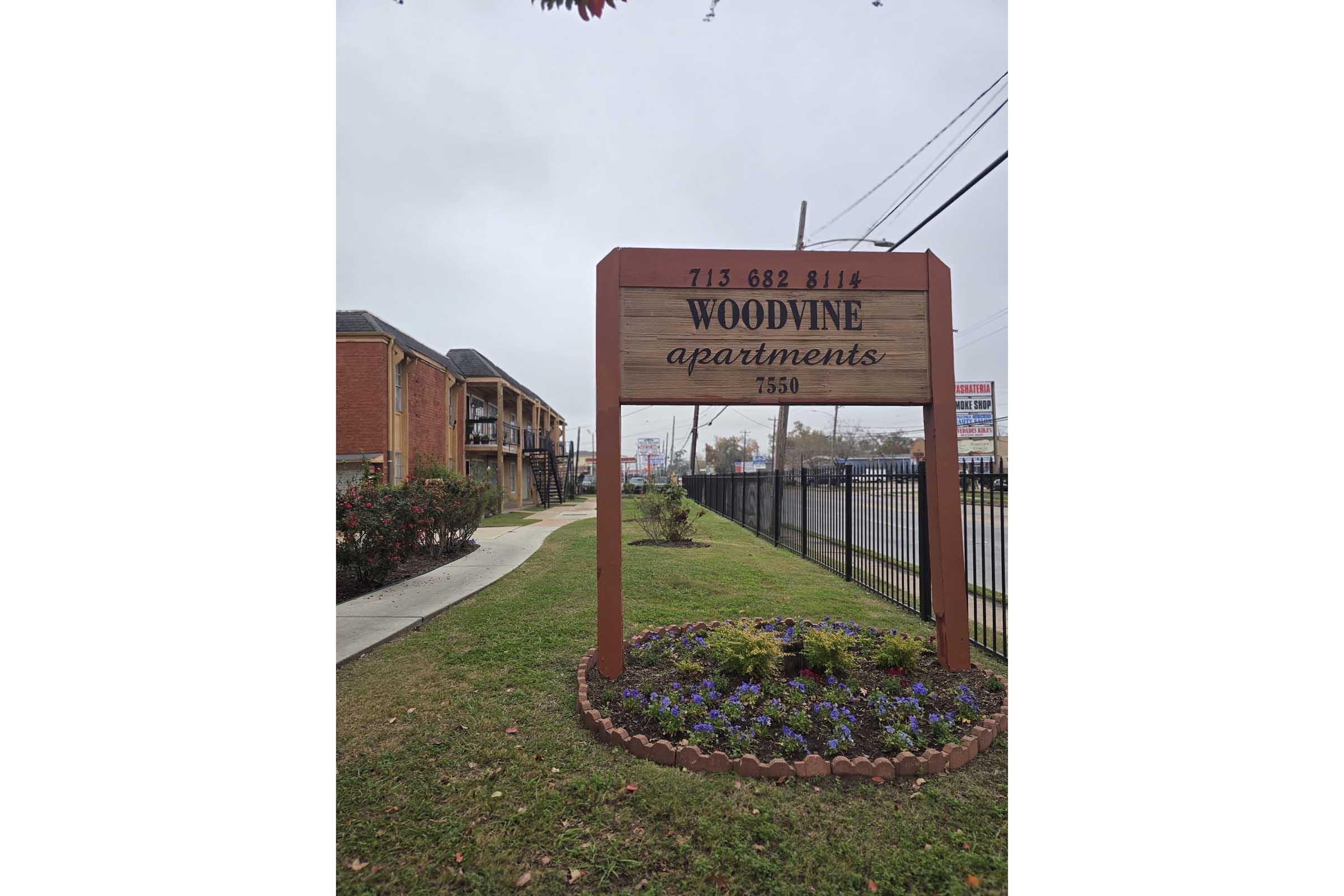 Sign for Woodvine Apartments located at 7550 with a phone number (713) 682-8114. The sign is framed in wood, surrounded by landscaped flowers and grass, with a cloudy sky in the background. The apartment complex is visible on the left side of the image.