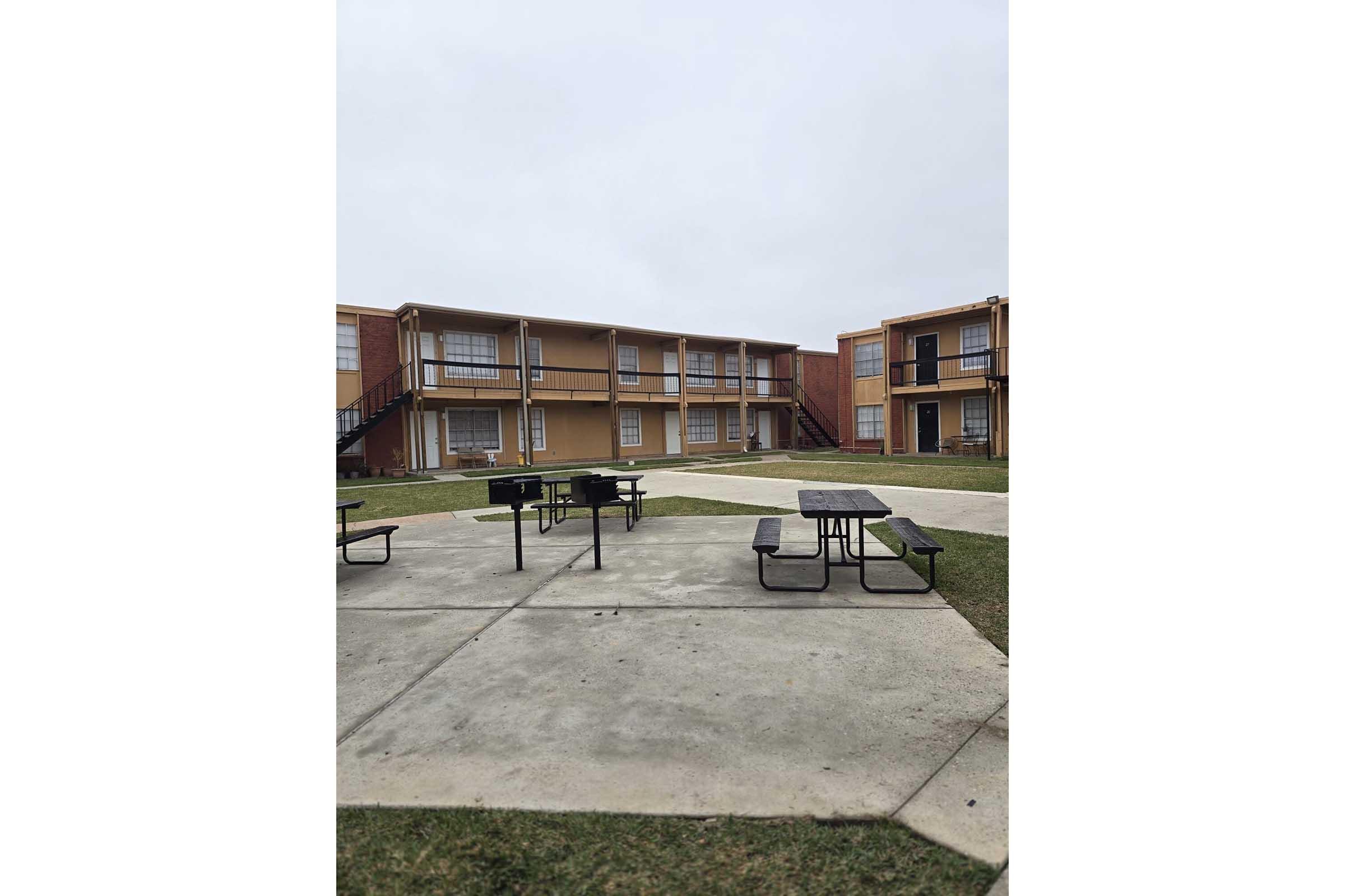 A view of a courtyard in an apartment complex featuring two-story buildings with balconies. In the foreground, there are several picnic tables on a concrete surface, surrounded by green grass. The sky is overcast, suggesting a cloudy day.
