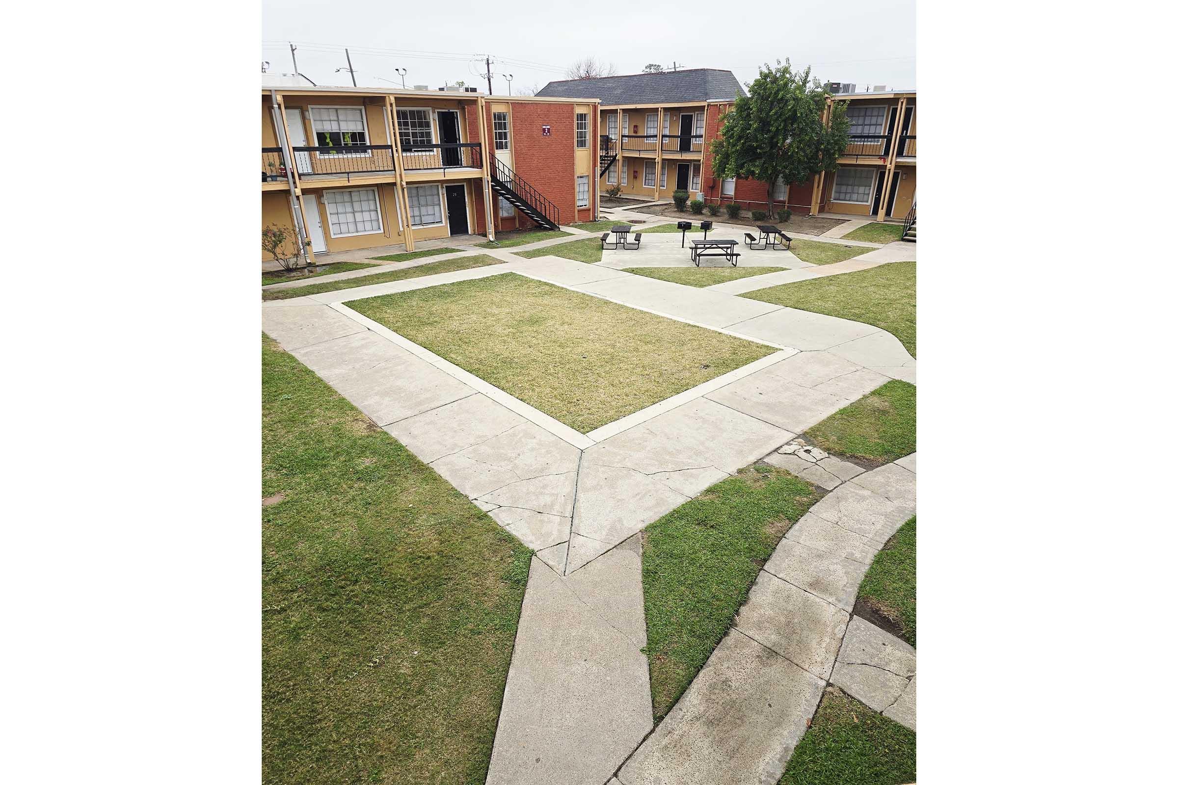 A view of an outdoor courtyard in an apartment complex featuring grassy areas and concrete pathways. The pathways intersect and lead to various apartment buildings, with benches visible in the center. The scene is cloudy and lacks people, creating a calm, residential atmosphere.