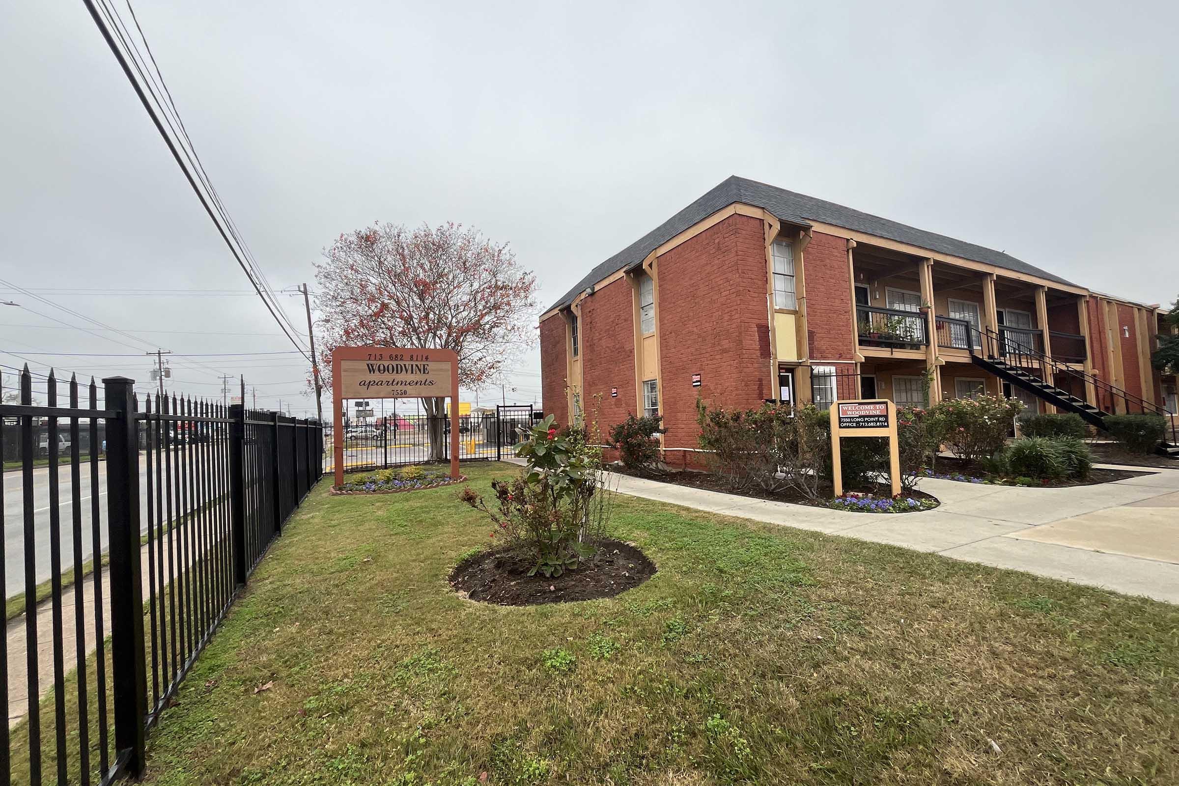 View of the Wooding Apartments, a two-story brick building with balconies, surrounded by a landscaped area featuring a small garden and signage. A fenced area borders the property, with a view of a street in the background under a cloudy sky.