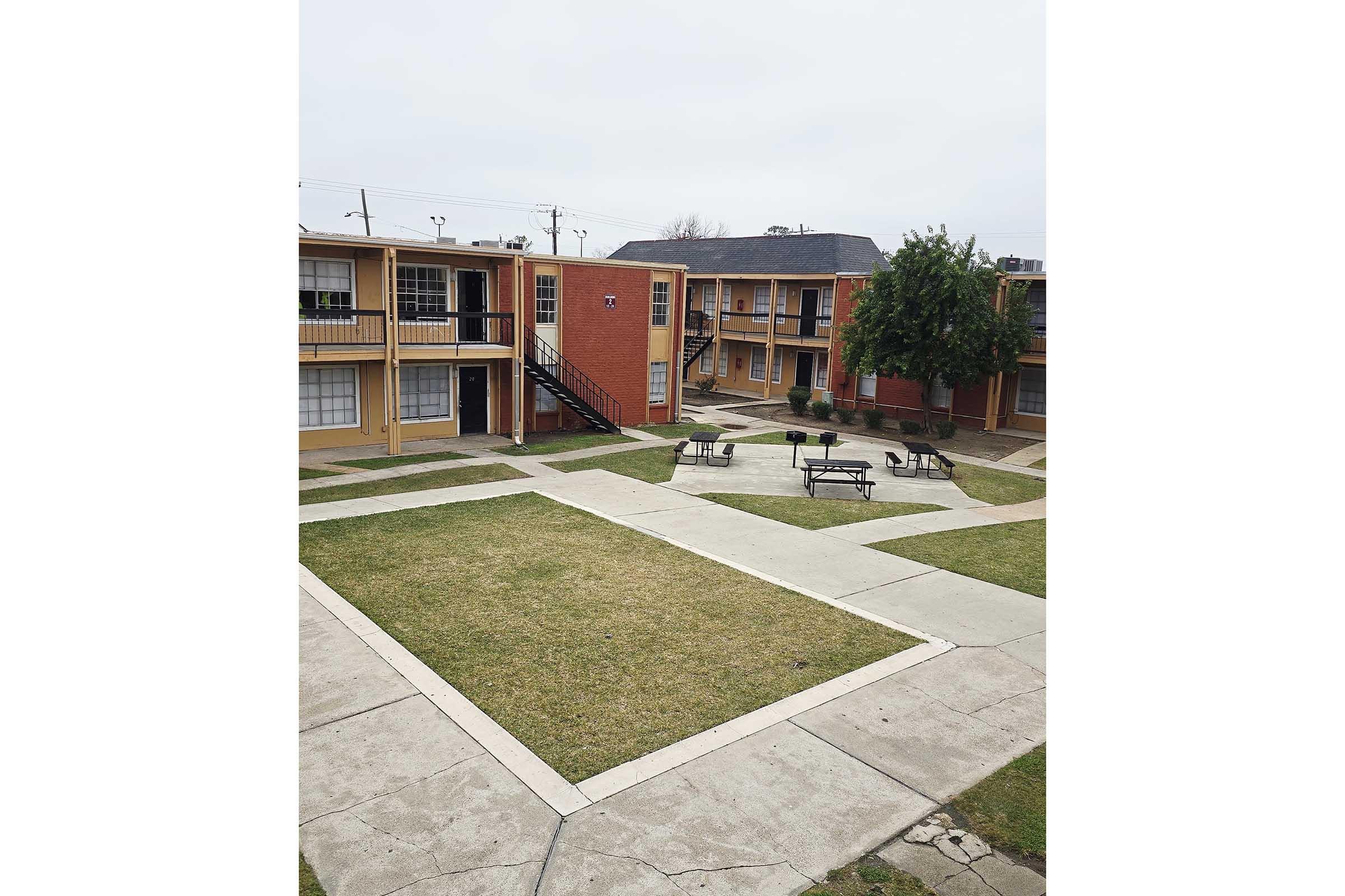 A view of a small apartment complex with two-story buildings arranged around a central courtyard. The courtyard features green grass, concrete pathways, and benches, with trees and overcast skies in the background.