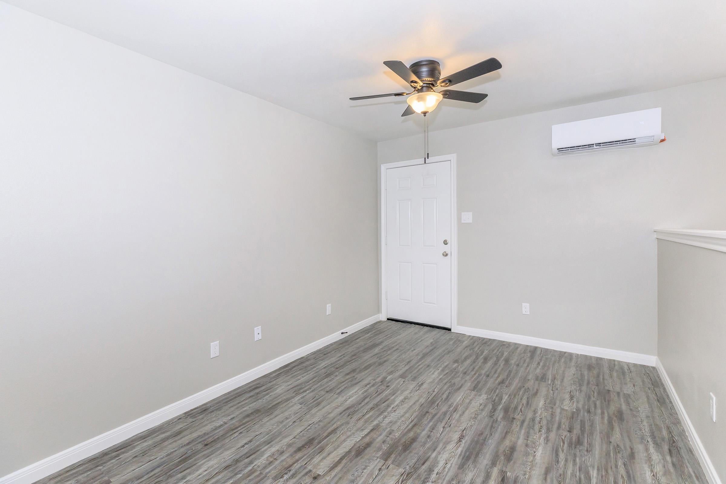 A vacant room with light gray walls, a ceiling fan, and a white air conditioning unit. The floor features wood-like laminate, and there's a closed white door leading outside. The space is well-lit and empty, ideal for customization or moving in furniture.
