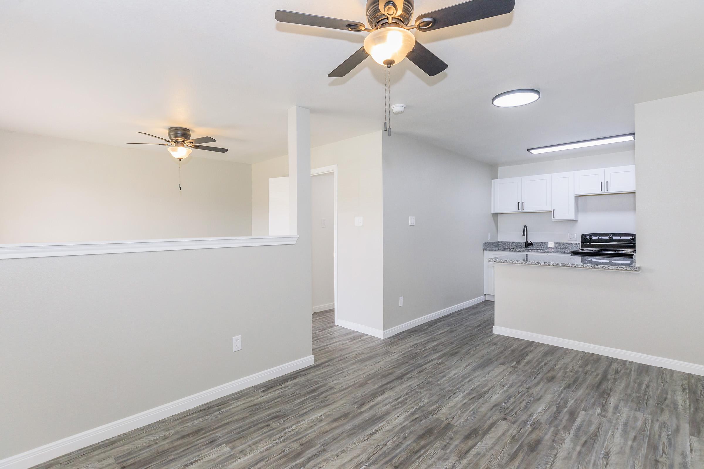 A spacious, modern apartment interior featuring light gray walls, a kitchen area with white cabinets, and a gray countertop. Two ceiling fans are installed, and there is a bright overhead light. The floor is covered in a stylish laminate resembling wood, enhancing the contemporary feel of the living space.