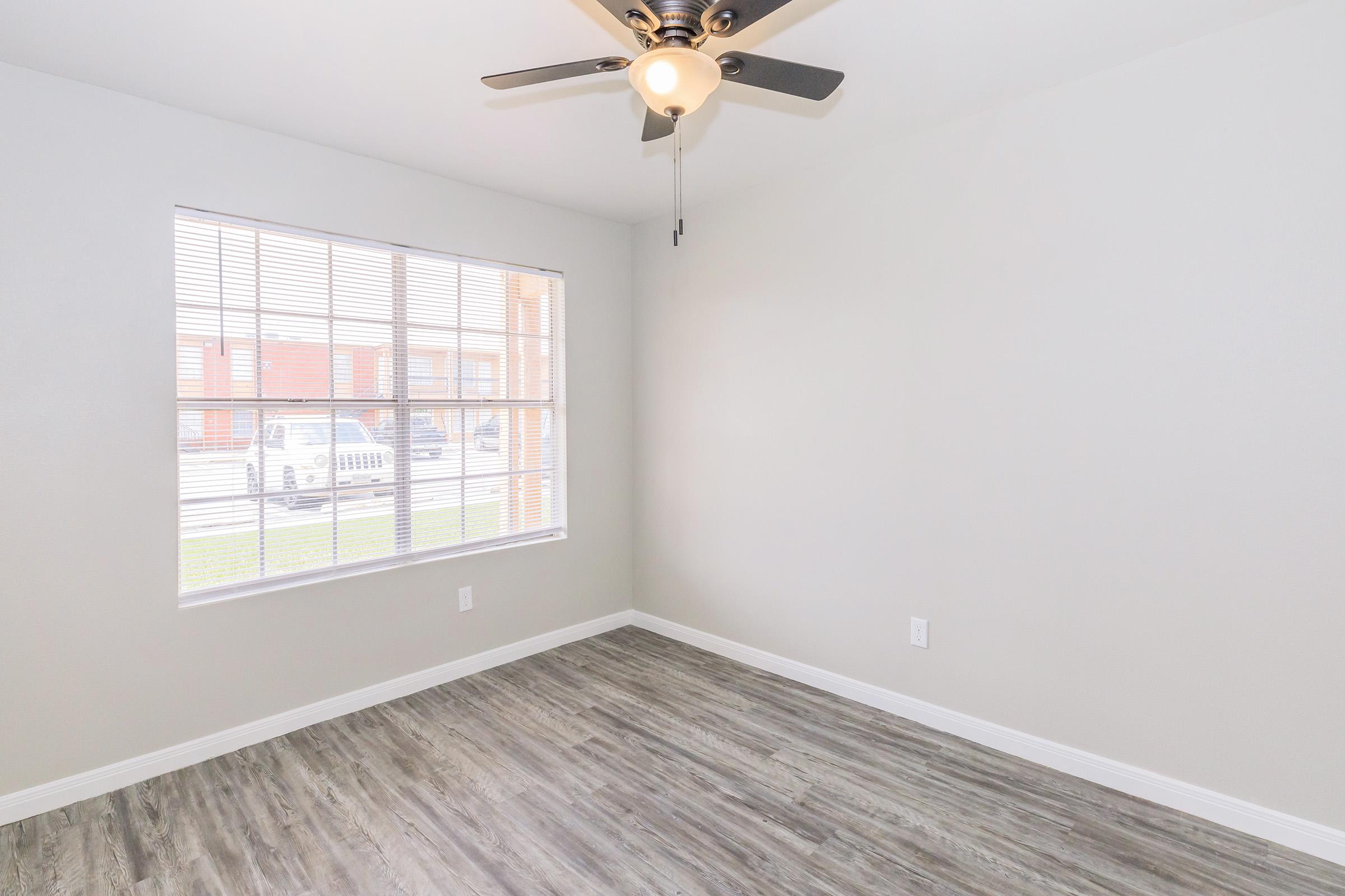 Empty room with light gray walls, a ceiling fan, and a large window with blinds. The flooring is a wood-like laminate, and the window overlooks an exterior view. The space appears bright and inviting, suitable for various interior designs.