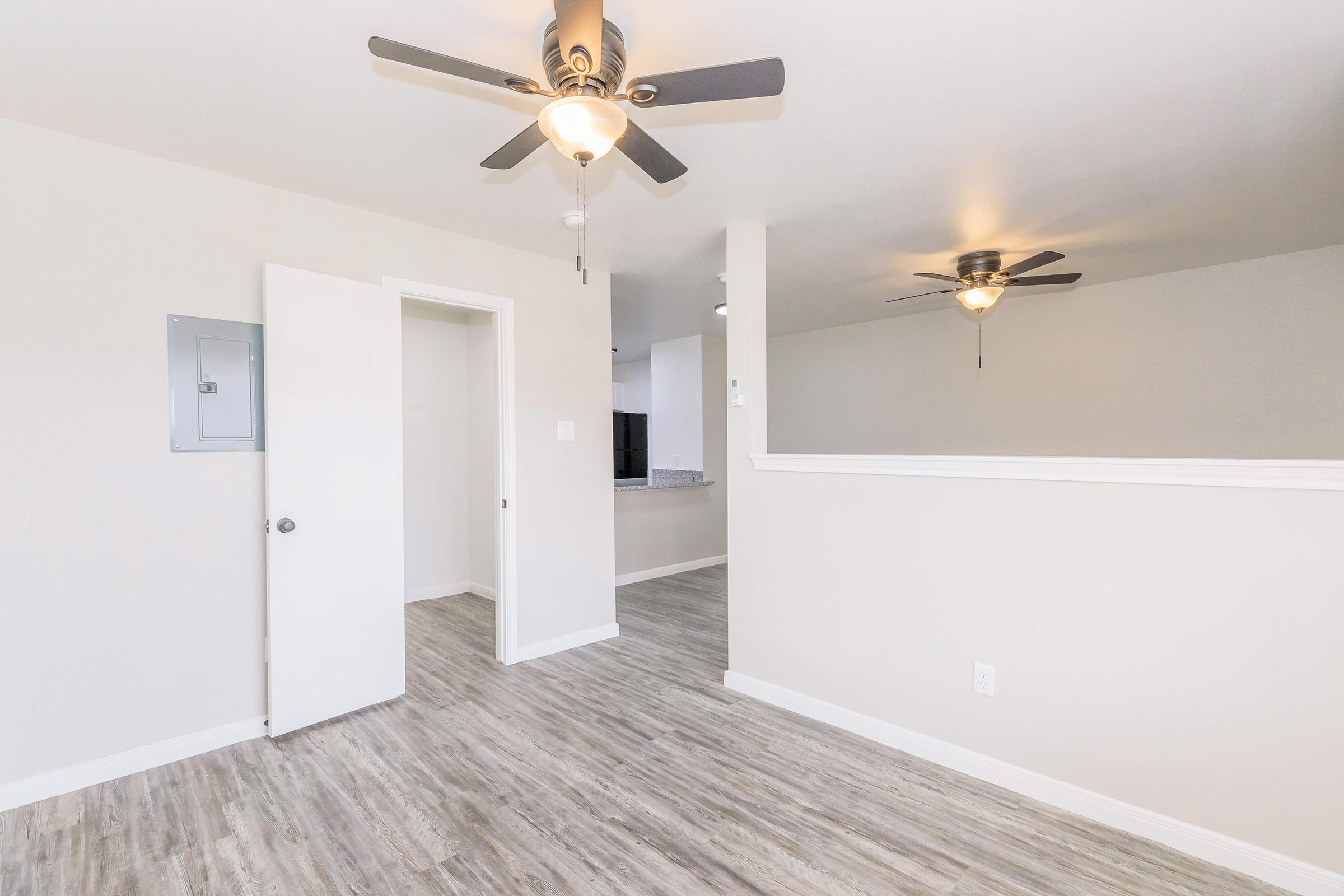An interior view of a room featuring light-colored walls and modern, wooden flooring. Two ceiling fans are visible, one in the foreground and one further back. A doorway leads to another room, and the setting appears bright and spacious.