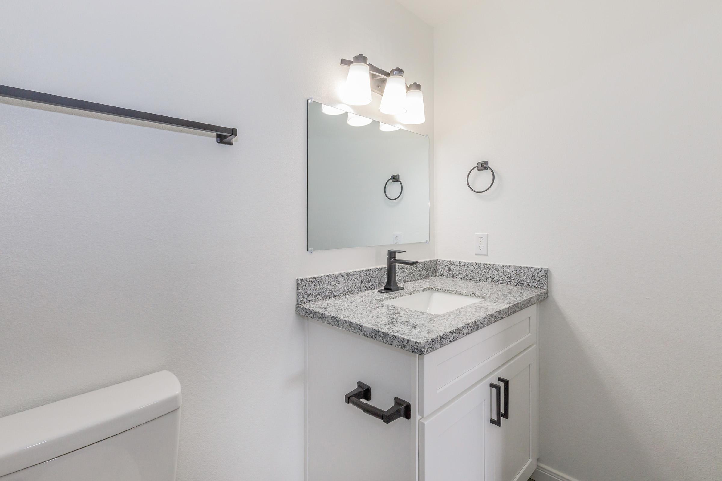 A modern bathroom with a white wall, featuring a granite countertop sink with a black faucet. A large mirror above the sink is illuminated by three light fixtures. There is a toilet on the left and a black towel bar mounted on the wall. The overall design is clean and minimalistic.