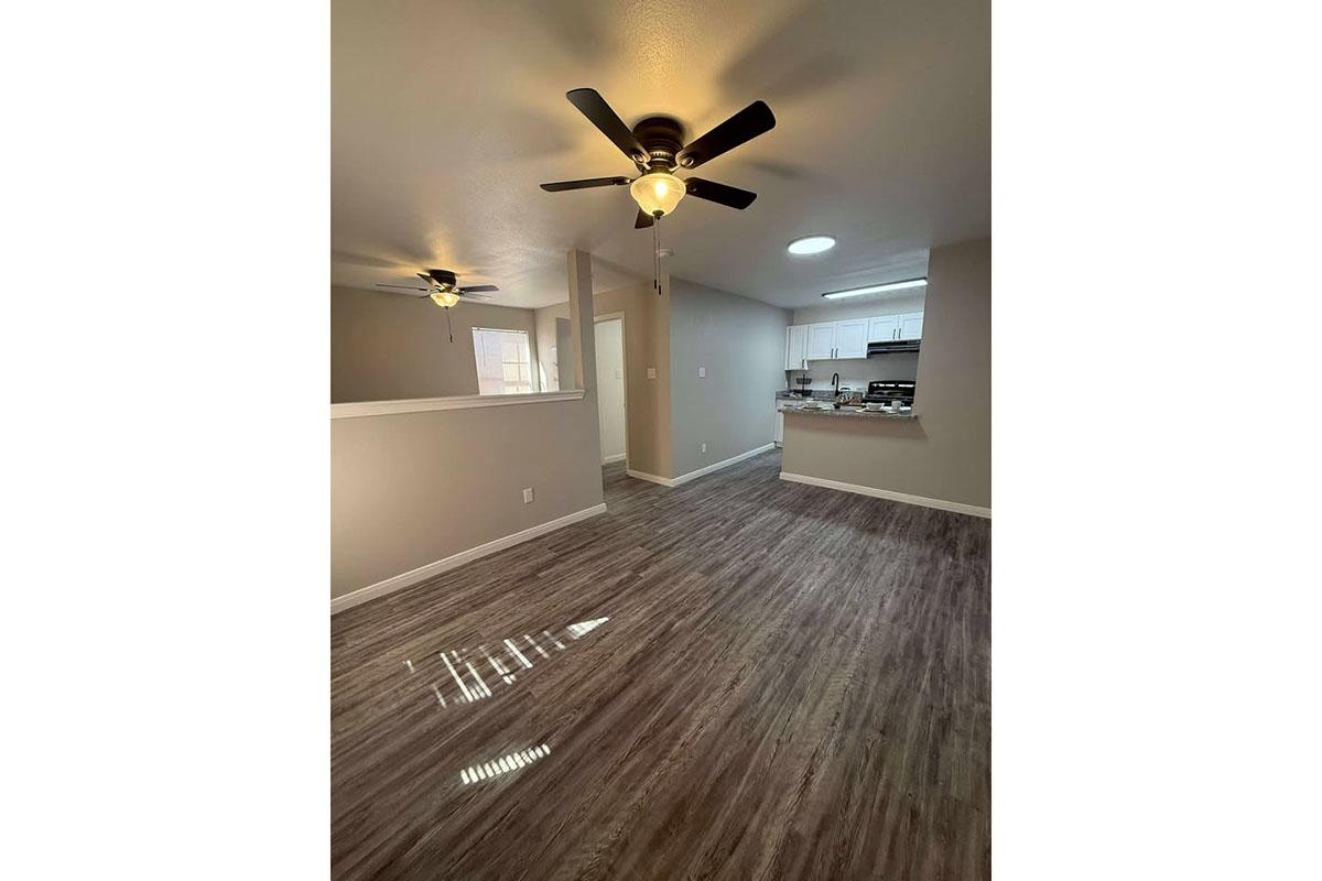 A spacious interior view of a well-lit room featuring a ceiling fan and laminate flooring. The room connects to a kitchen area visible in the background. The walls are painted in a neutral color, creating a bright and inviting atmosphere.