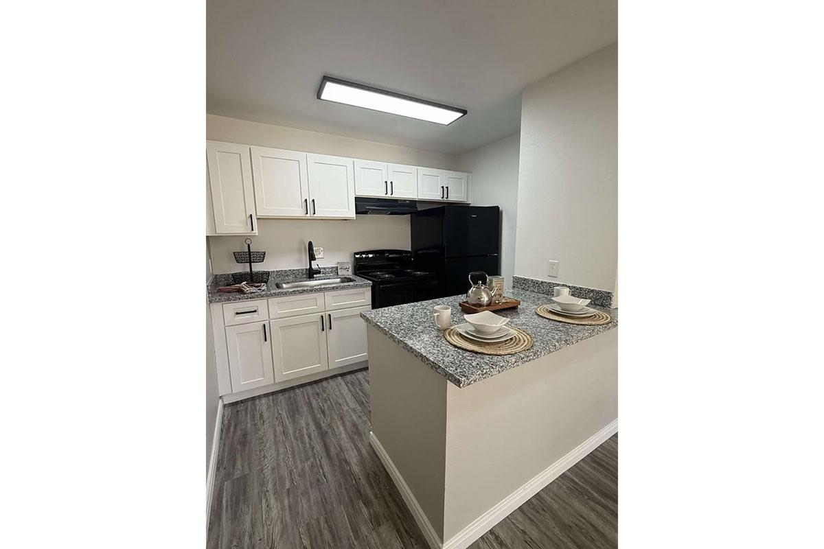 Modern kitchen featuring white cabinetry, a black stove and refrigerator, and granite countertops. A kitchen island with place settings for two is visible, showcasing plates and utensils. The flooring is a light wood finish, and there is ample overhead lighting.
