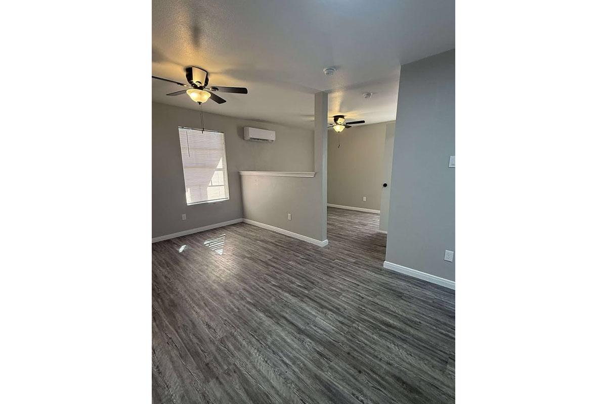 Interior of a modern room featuring gray walls, hardwood-style flooring, and ceiling fans. Sunlight streams in through a window, illuminating the space. A doorway leads to another room, indicating an open layout.