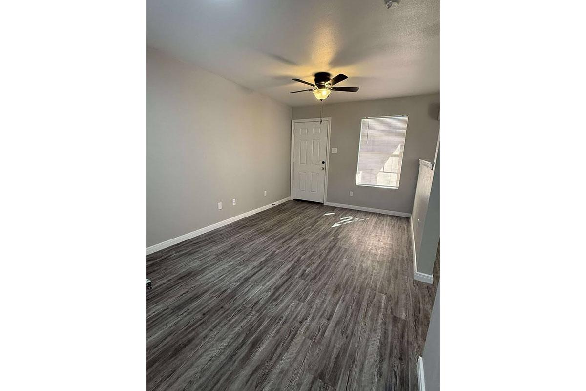 A spacious, well-lit living area with laminated flooring, a ceiling fan, and a front door. The walls are painted in a light color, and a small window allows natural light to enter. The room is empty, creating a blank canvas for furniture and decoration.