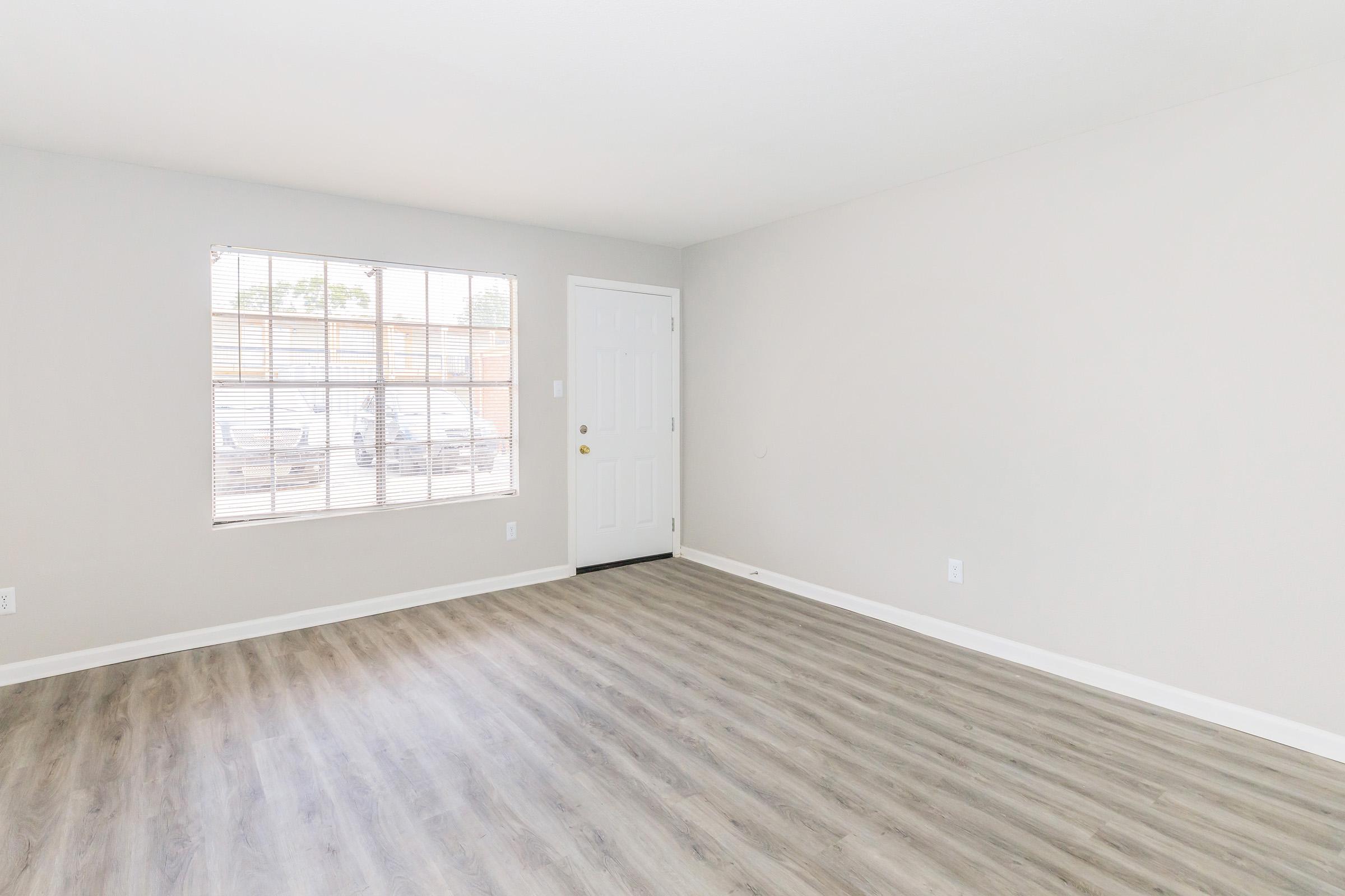 A spacious, empty room with light gray walls and laminate flooring. There is a large window with white blinds on one wall, allowing natural light to enter. The door is a simple white design, and the overall ambiance is bright and airy, suitable for various interior setups.