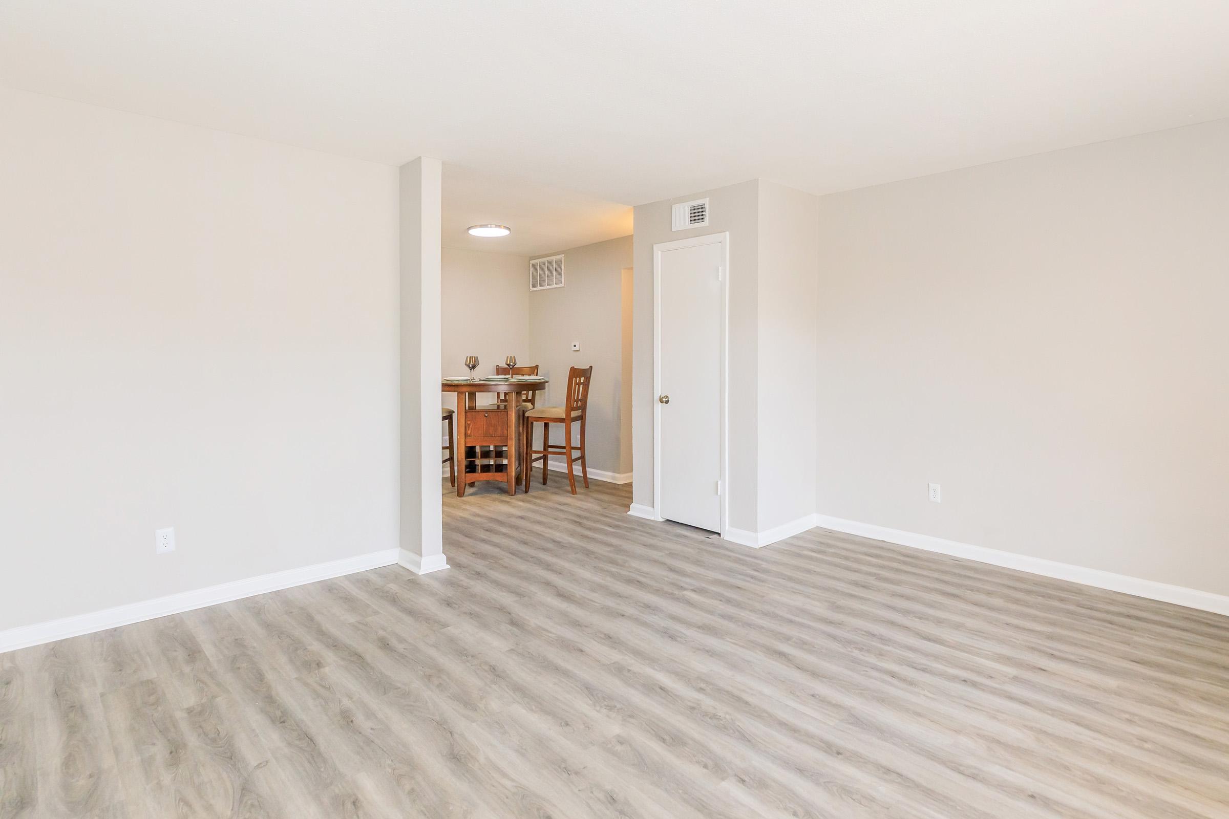 A bright and airy interior of an empty room featuring light-colored walls and light wood laminate flooring. To the right, there is a small dining area with a table and chairs, and a door leading to another space. The room has ample natural light, creating an inviting atmosphere.