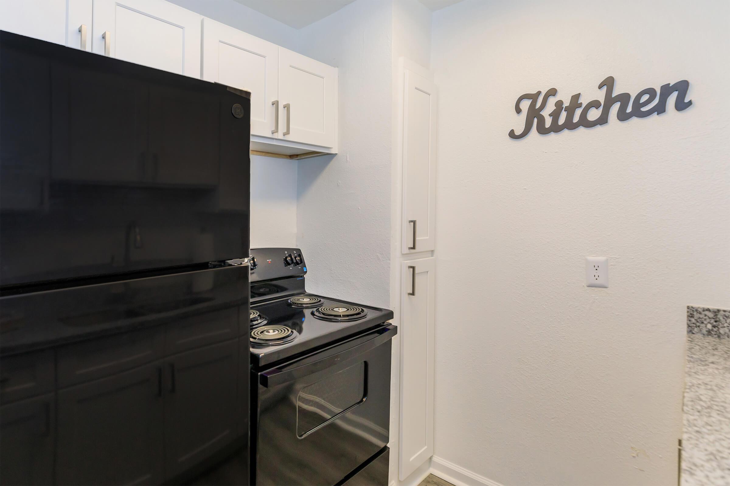 A modern kitchen featuring a black refrigerator and stove with four burners. The cabinets are white, and there is a decorative sign on the wall that reads "Kitchen." The countertop has a light gray finish, complementing the overall sleek design of the space.