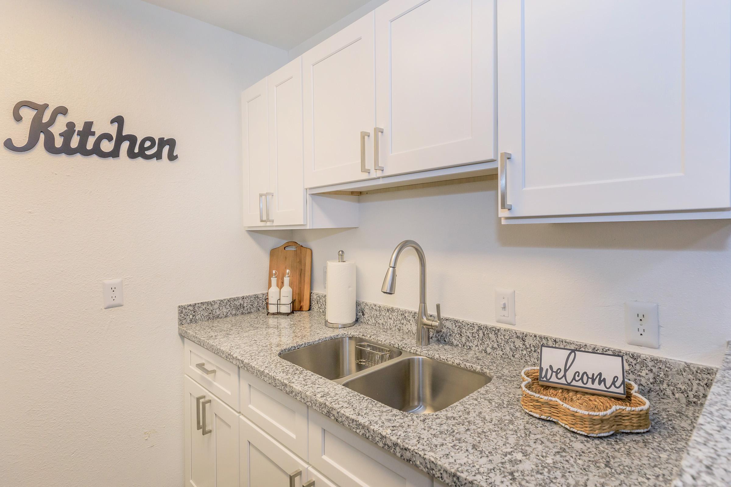 Modern kitchen featuring white cabinets, a granite countertop, and a double sink. A wooden cutting board and bottles are neatly arranged beside the sink. A “welcome” sign is placed on the counter, and the word "Kitchen" is displayed on the wall. The overall décor is clean and inviting.