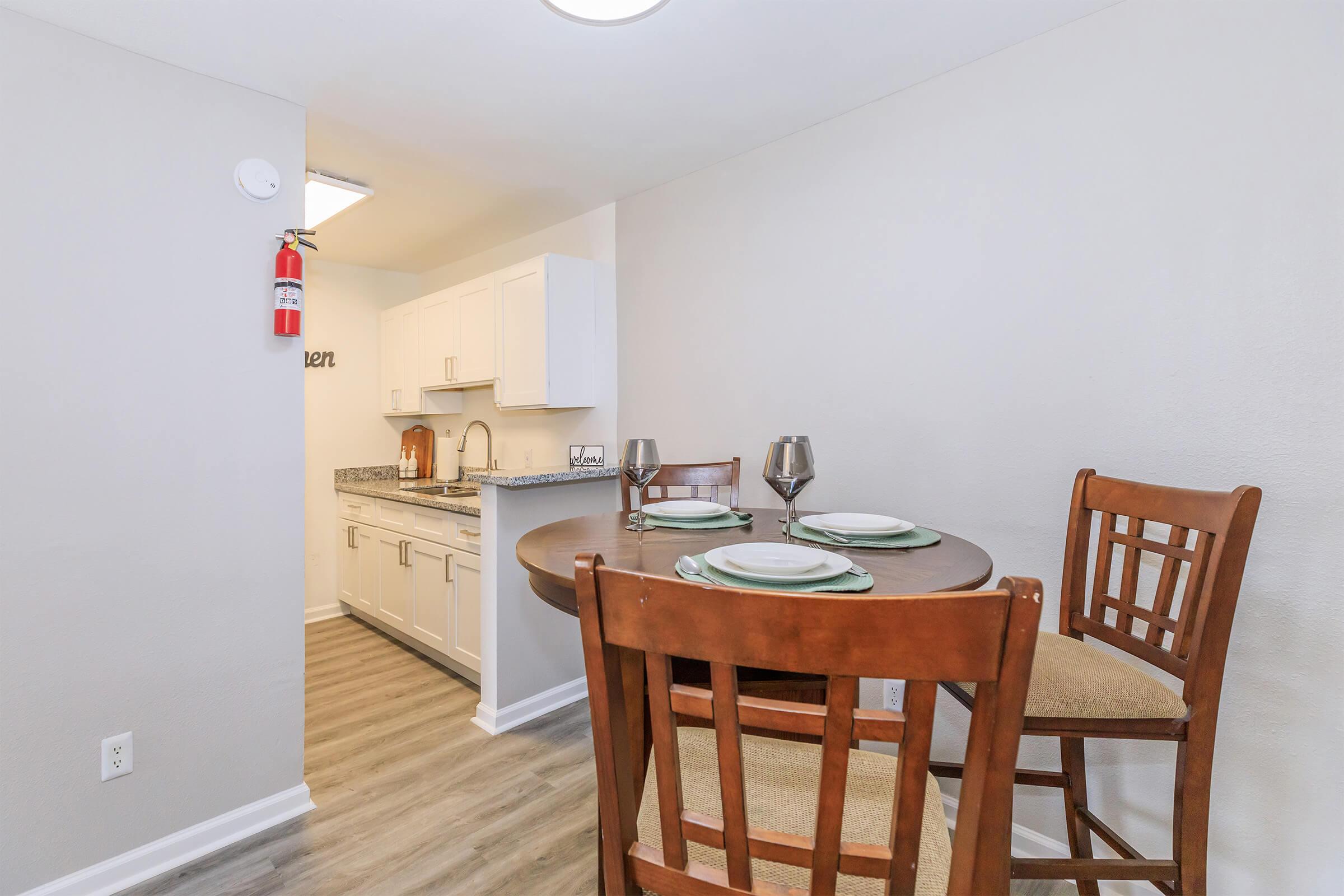 A cozy dining area features a round table set for two, with dishes and silverware. In the background, a modern kitchen is visible, showcasing white cabinetry and a granite countertop. Soft, neutral wall colors and elegant lighting create a welcoming atmosphere.