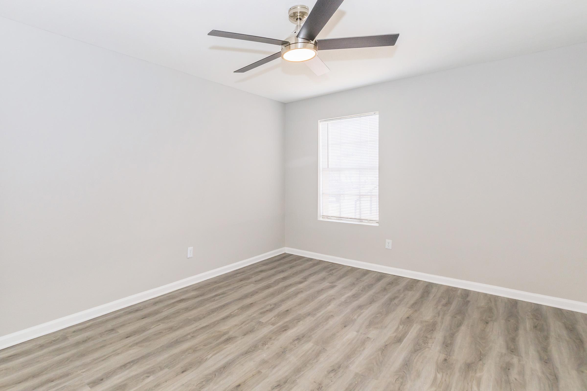 A simple, empty room with light gray walls and wood-style flooring. The space features a ceiling fan with a circular light fixture and a window on the far wall allowing natural light. The overall atmosphere is bright and airy, reflecting a minimalist design.