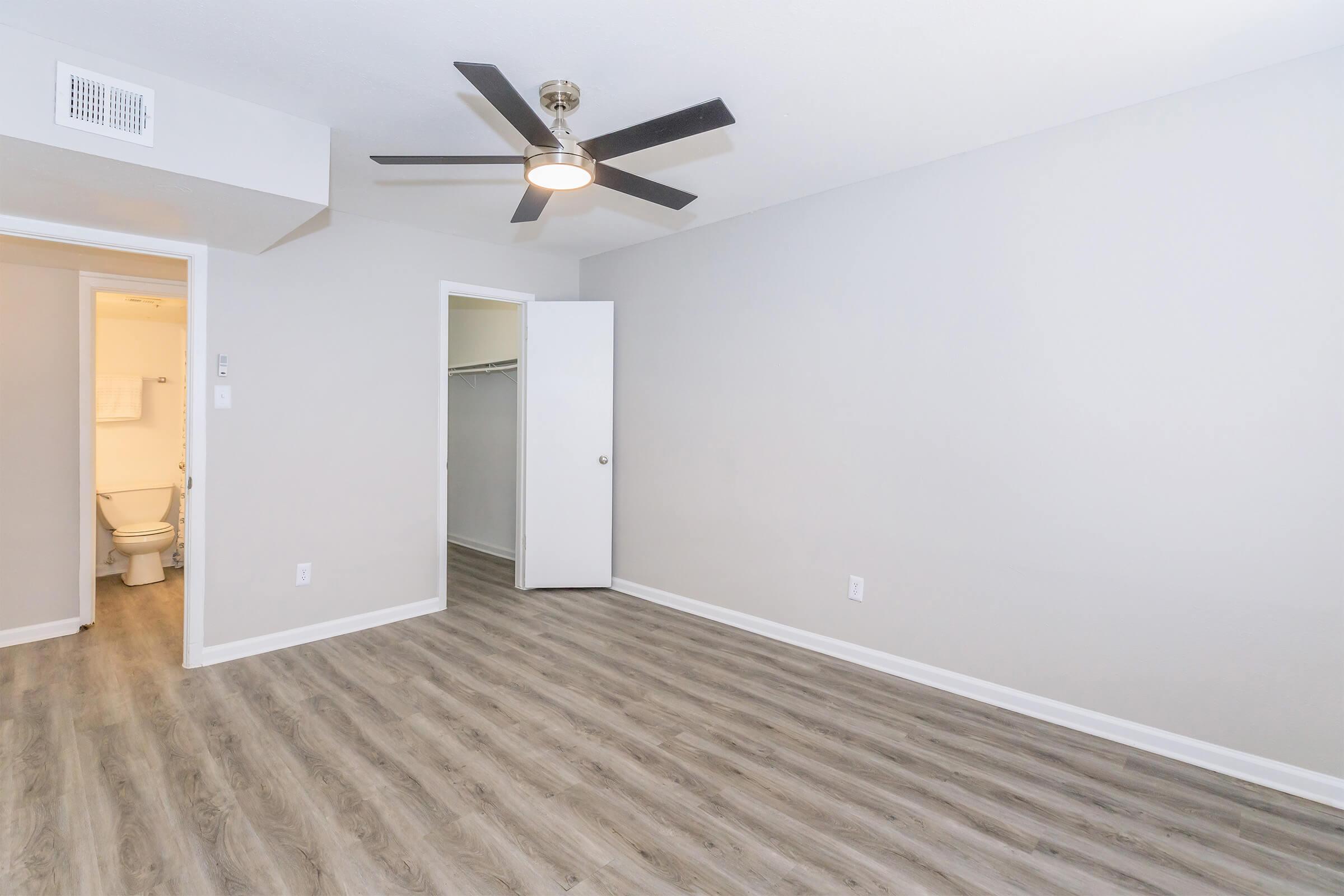 Empty room with light gray walls and light wood-like flooring. A ceiling fan is visible, and there's a doorway leading to a bathroom area with a toilet. The room has a spacious feel, with no furniture present and a closet area on one side.