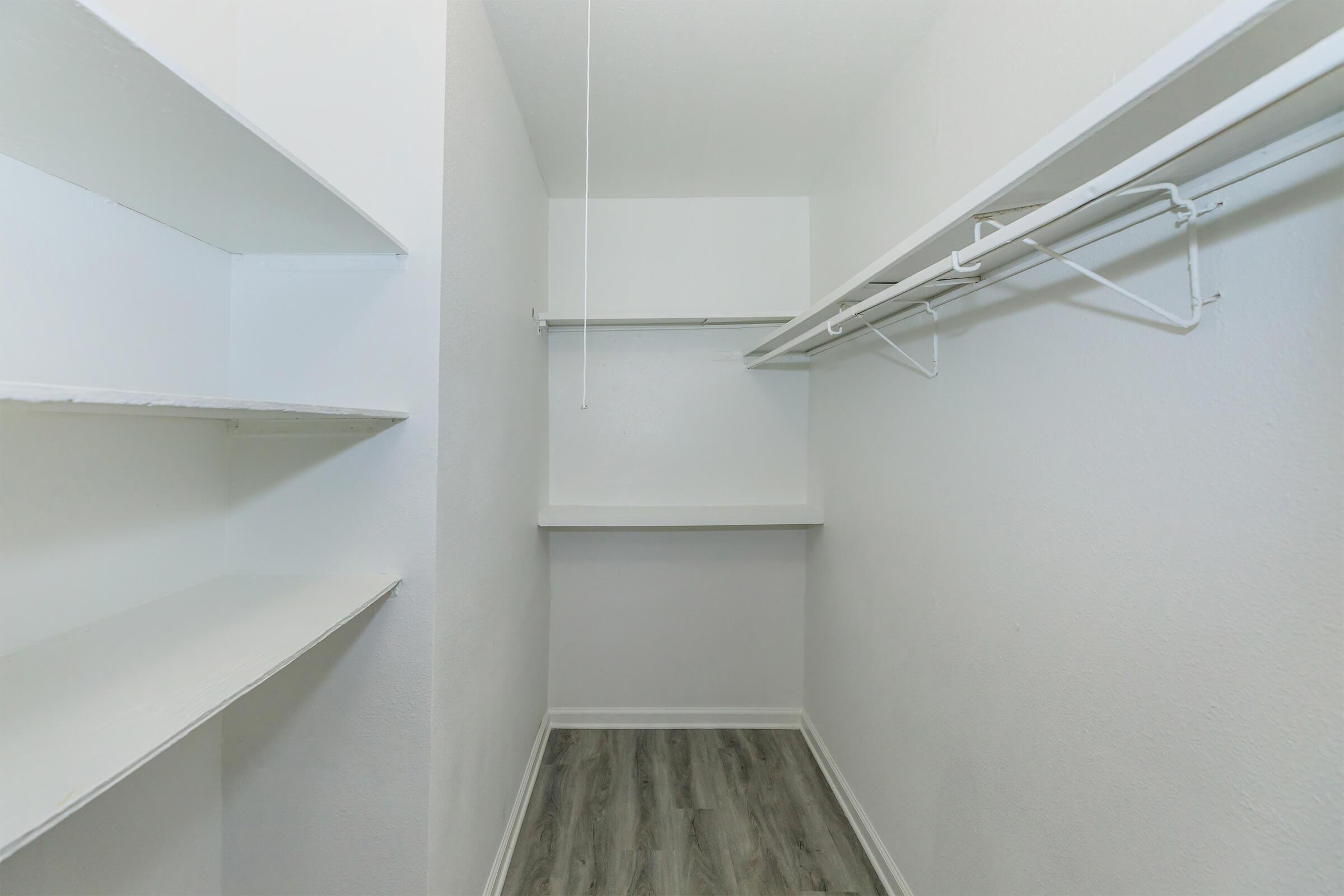 A spacious, empty closet featuring white walls and shelves on the left side. The floor is covered with light-colored laminate, and a hanging rod is installed above a flat surface. The closet has a minimalistic design, making it ideal for organizing clothes and accessories.