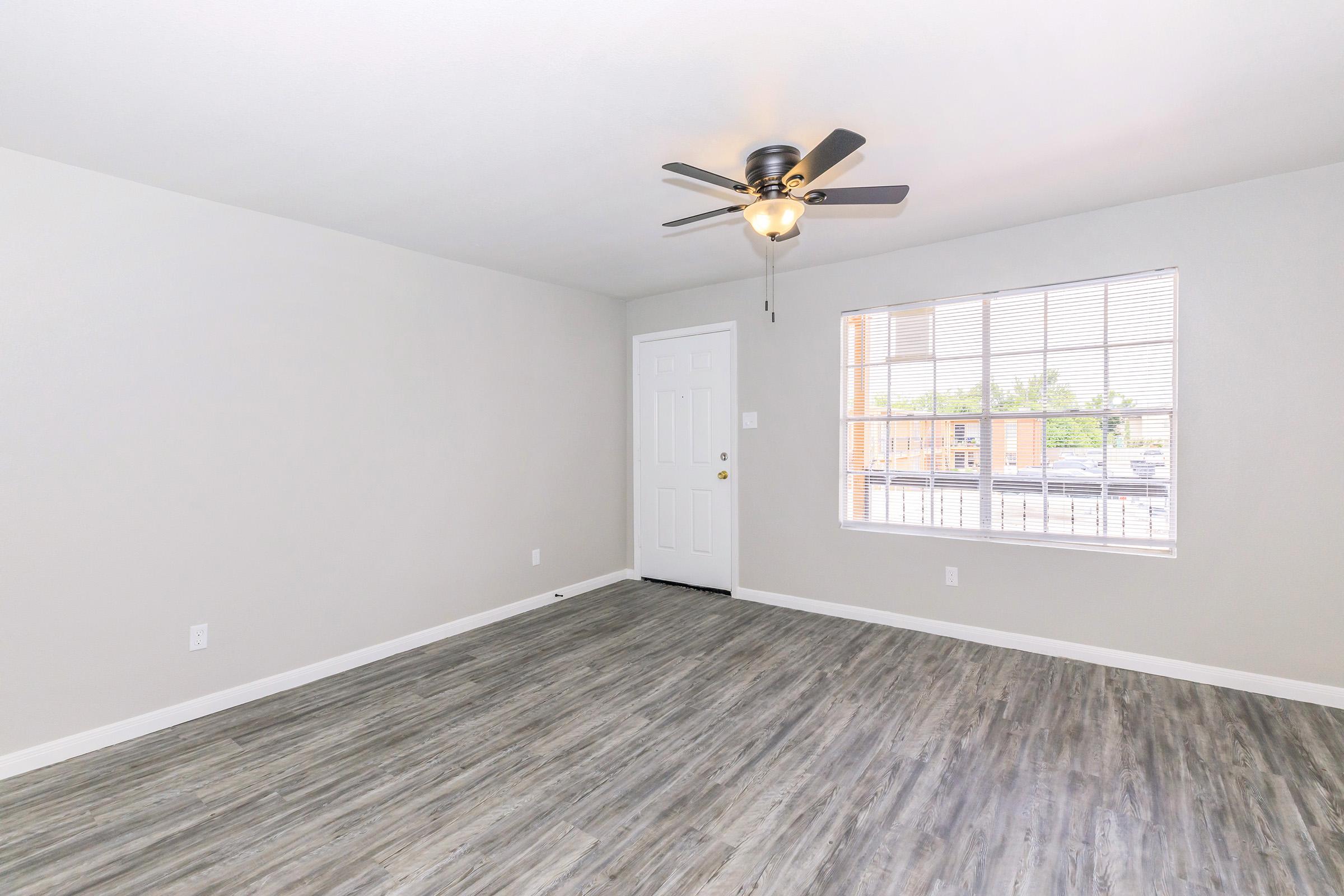 A spacious, empty room featuring light-colored walls and a ceiling fan. The floor has a wood-like finish. A large window with blinds allows natural light, and there is a door leading outside. The overall atmosphere is bright and inviting, perfect for personal decoration.