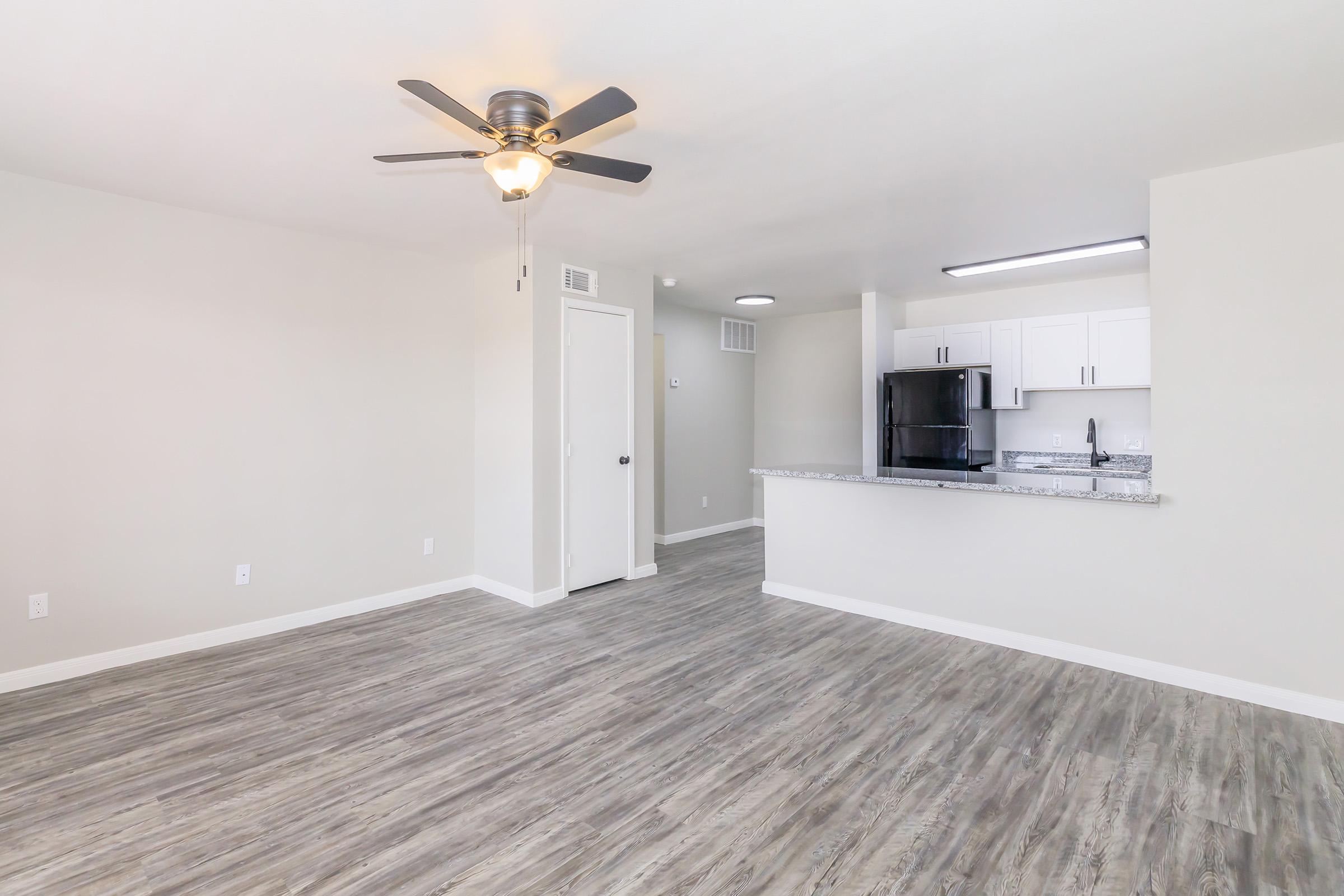 A bright, spacious living area featuring light-colored walls and sleek, wood-like flooring. A ceiling fan is installed in the center, while a modern kitchen with white cabinets and a granite countertop is visible in the background. The space is well-lit, showcasing an inviting atmosphere.