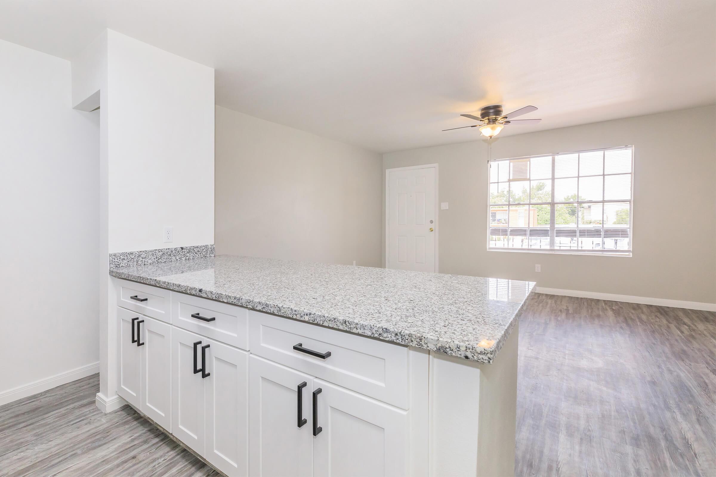 Interior view of a modern kitchen with a granite countertop and white cabinets. The open layout leads to a spacious living area with a large window and a ceiling fan. The flooring is a light grey, enhancing the bright and airy atmosphere of the space.