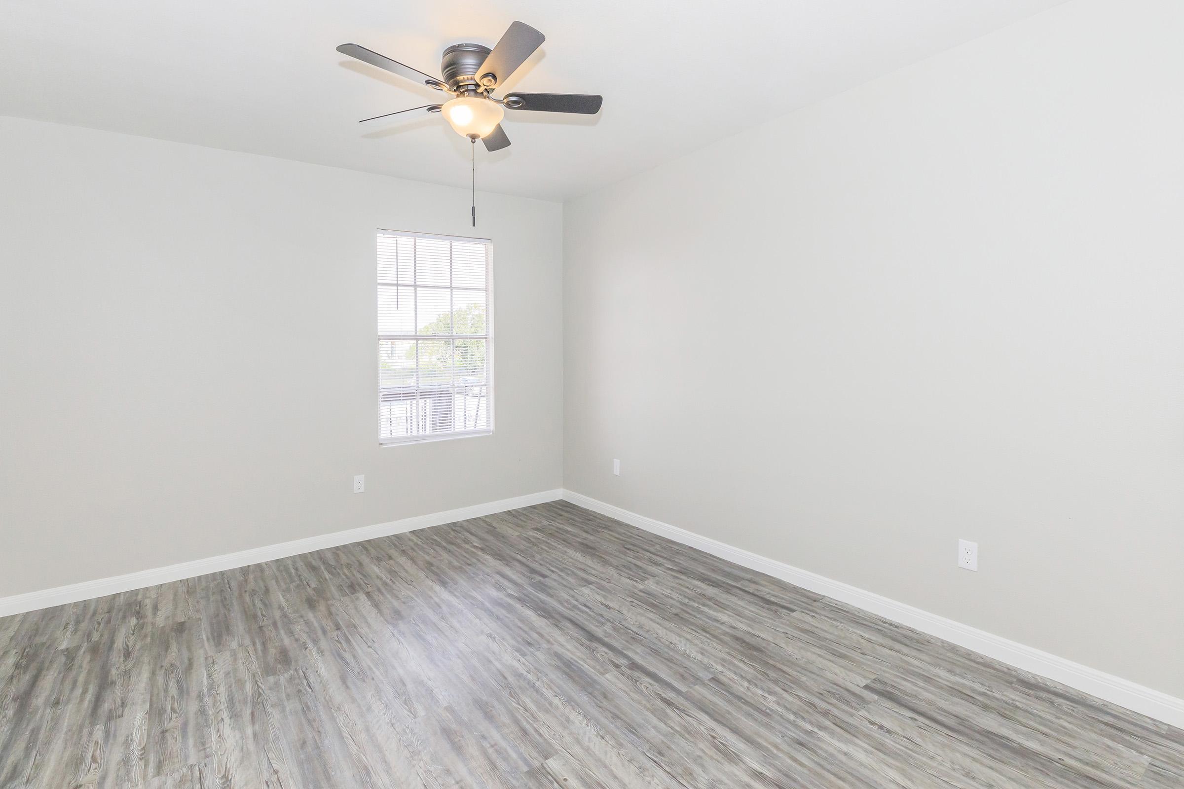 A well-lit, empty room featuring light gray walls, a ceiling fan, and a window with white blinds. The flooring is a wood-like laminate in warm tones. The space is spacious and ready for furnishing.